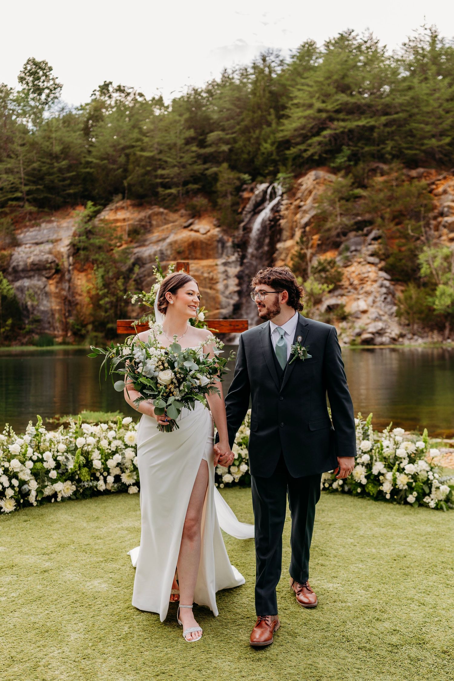 Smiling bride and groom hold hands at outdoor wedding ceremony with stunning waterfall backdrop.