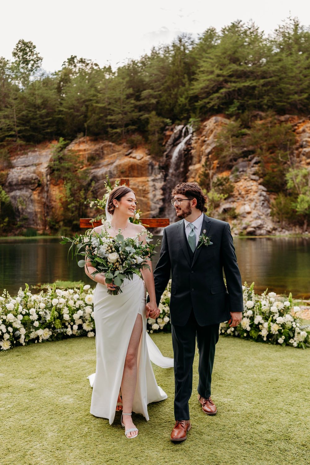 Smiling bride and groom hold hands at outdoor wedding ceremony with stunning waterfall backdrop.
