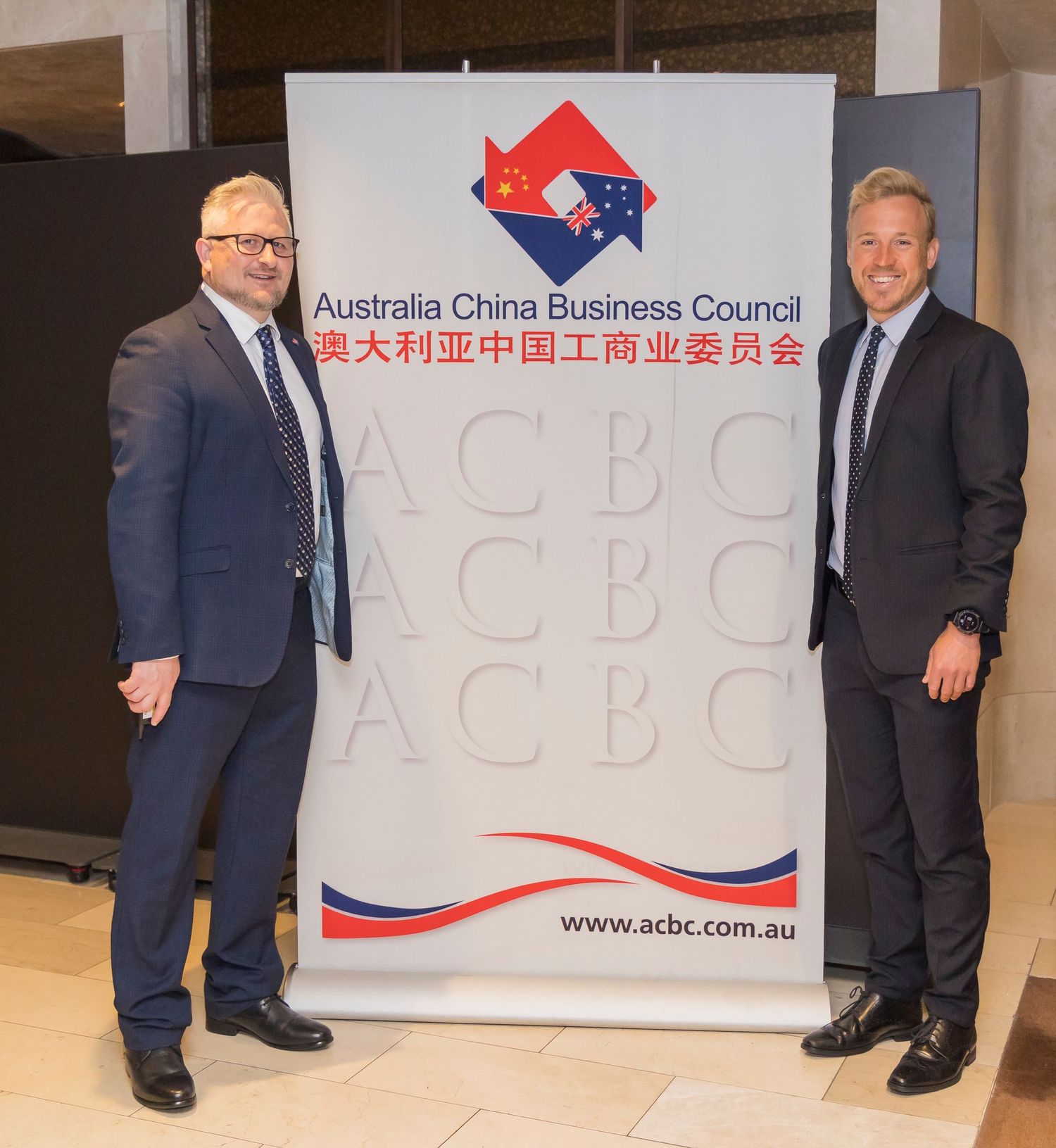 Two men in suits standing beside Australia China Business Council banner display.
