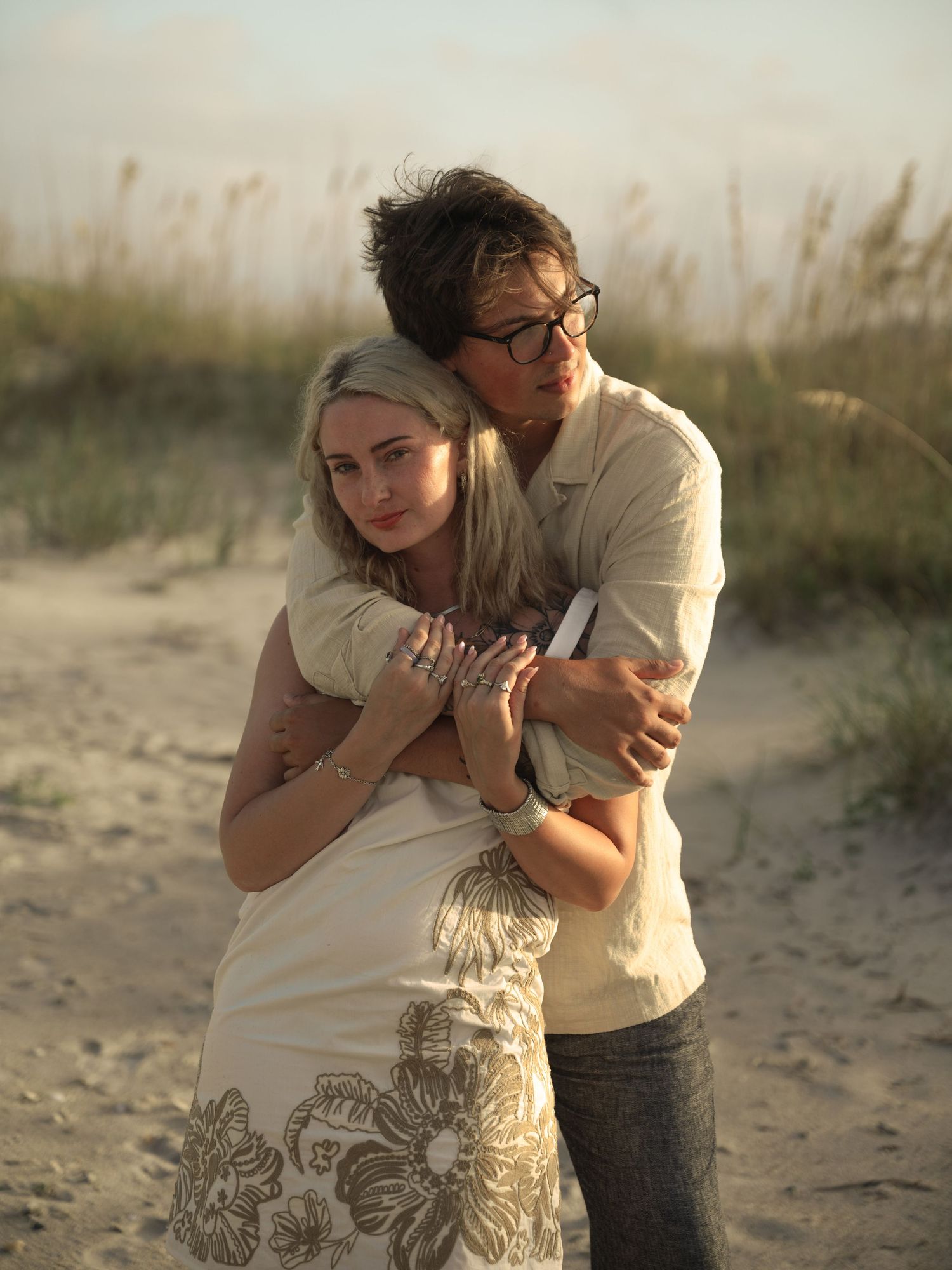 Romantic couple embracing on sandy beach at golden hour with tall grass in background.