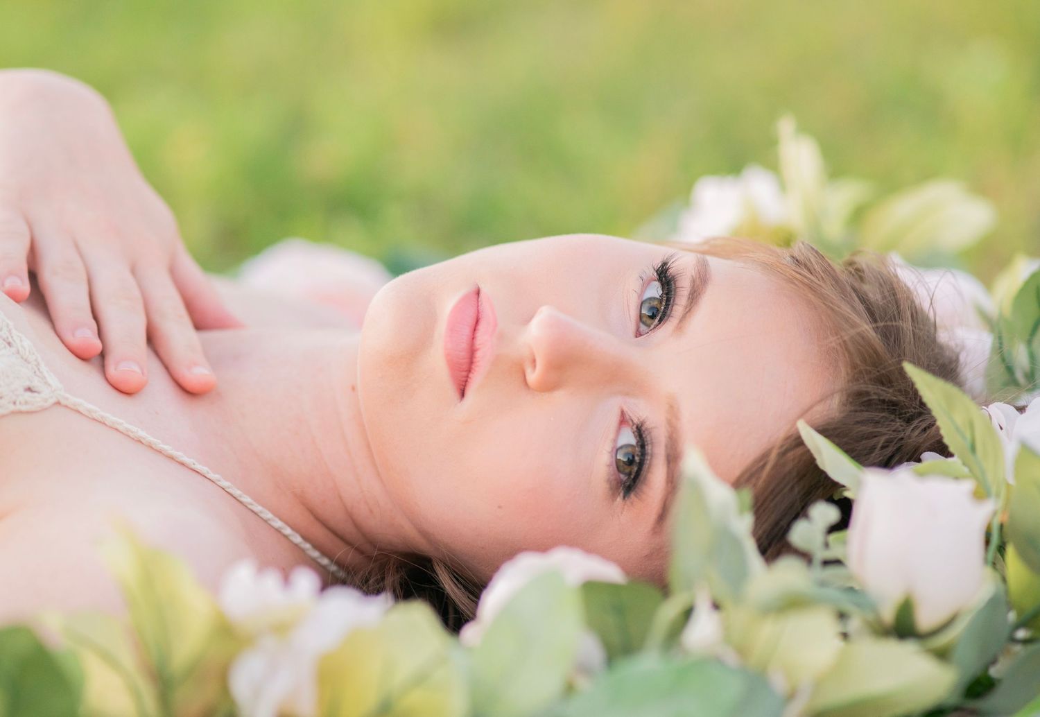 A young woman with blue eyes lies among white flowers in a green field, gazing dreamily at the camera.