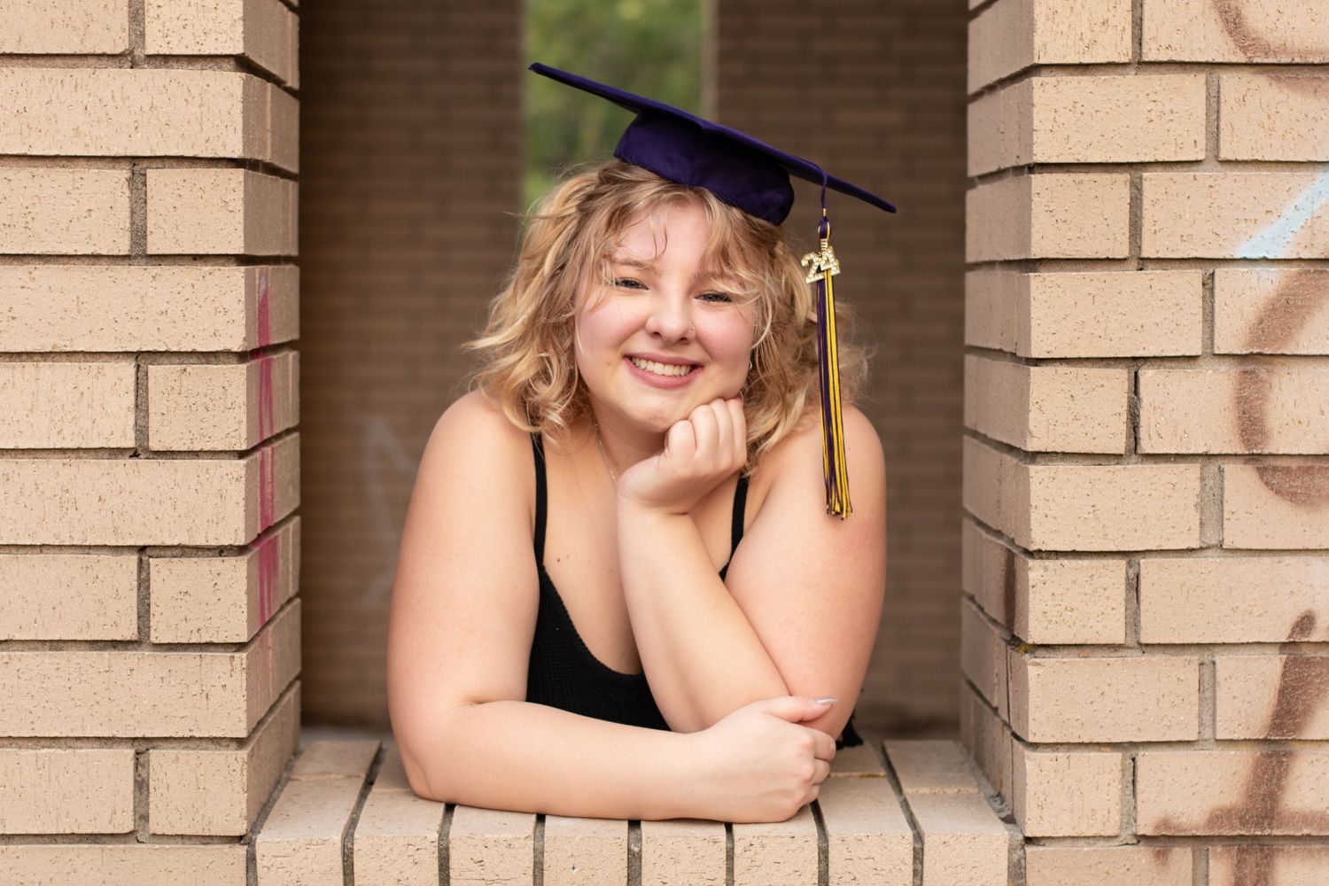 Graduate in black cap posing in brick archway during senior photo session.