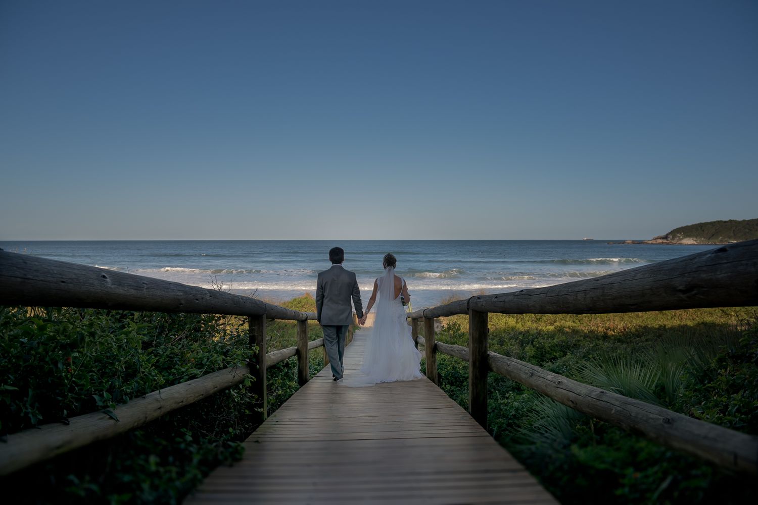 A couple stands on a wooden boardwalk overlooking a scenic beach at sunset.