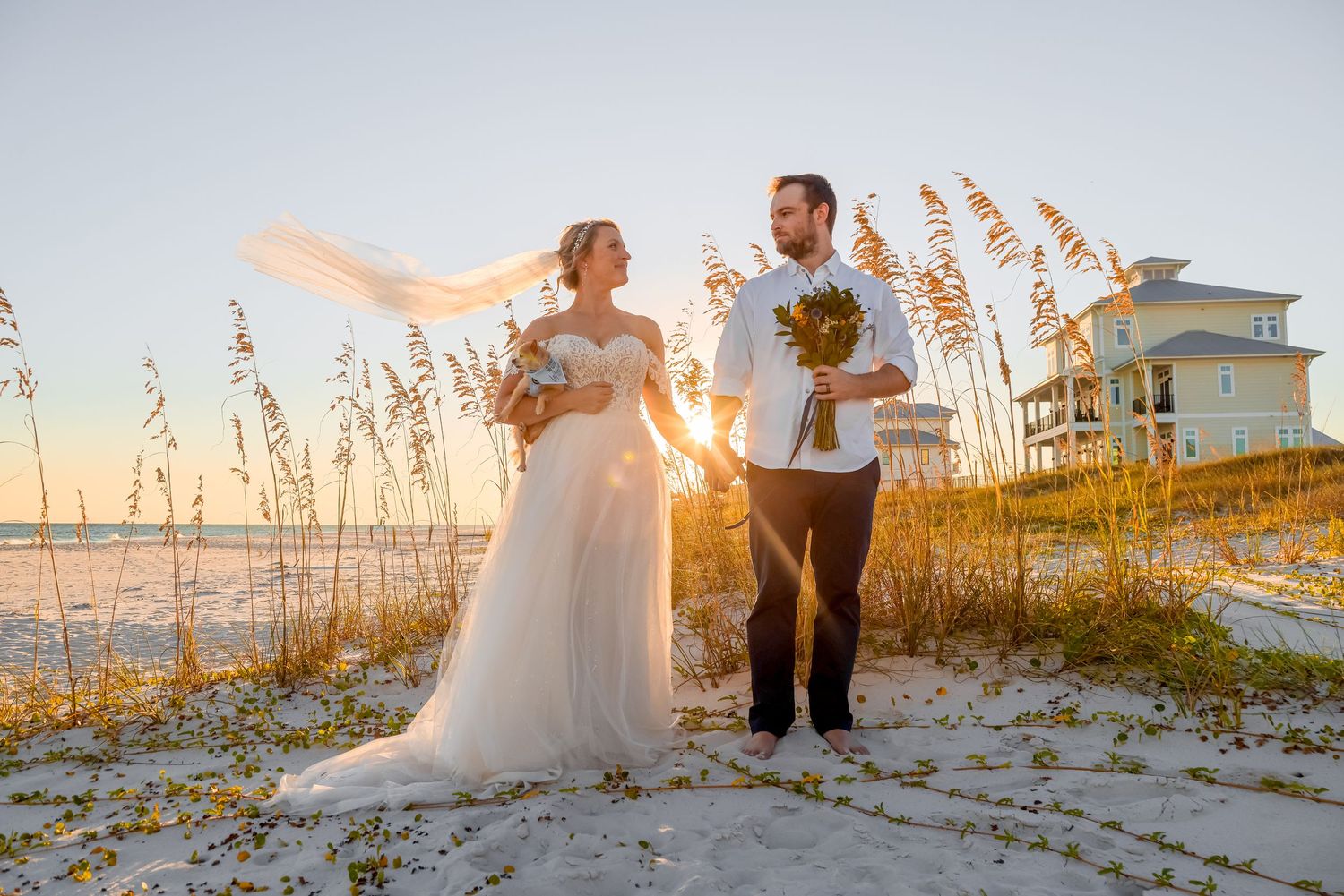 Newlyweds share a romantic moment at sunset on a sandy beach with sea oats in the foreground.