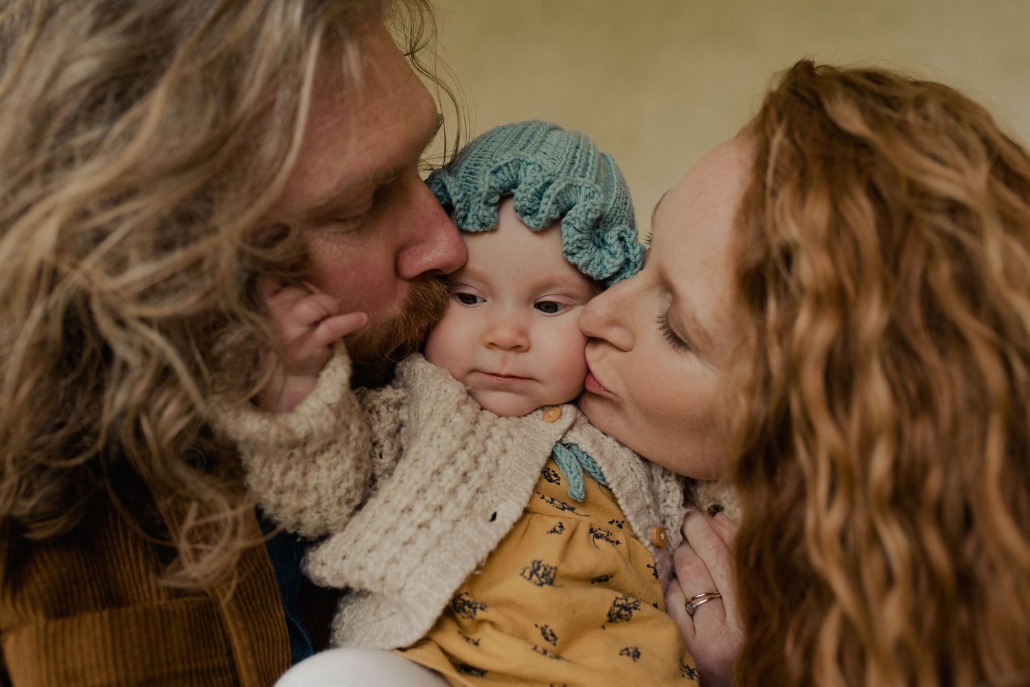 A baby wearing a blue bonnet receives kisses on both cheeks in a warm intimate moment.