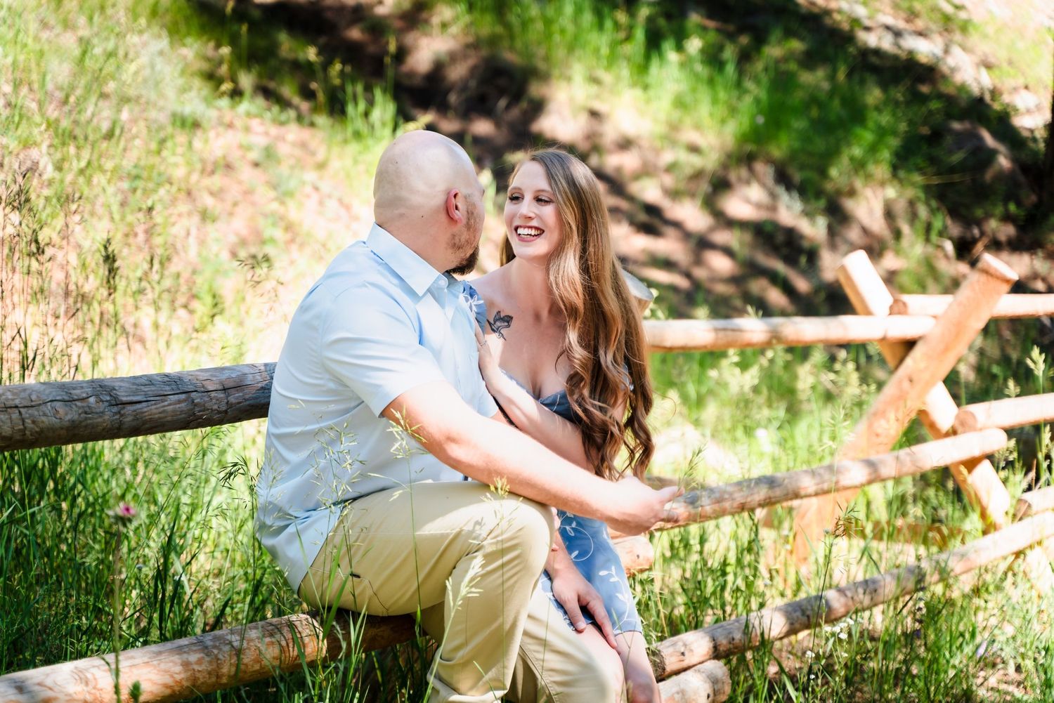 A romantic couple sits together on a wooden fence sharing a joyful laugh in a sunlit outdoor setting.