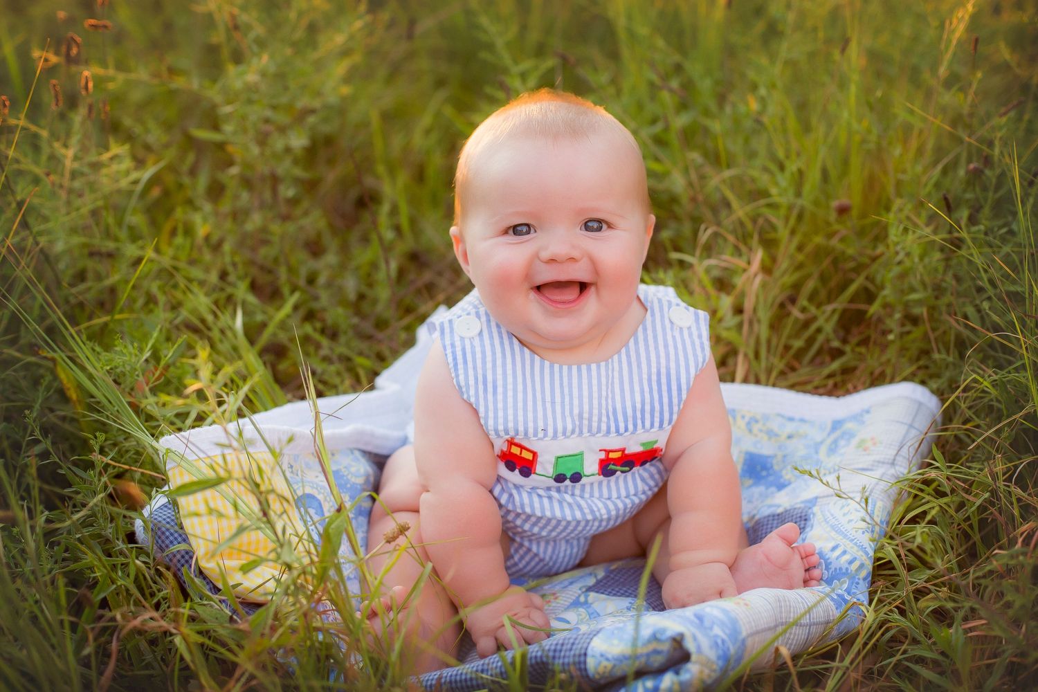 Six month old baby boy sitting in a green field at sunset.  Wearing a blue and white striped romper with trains.
