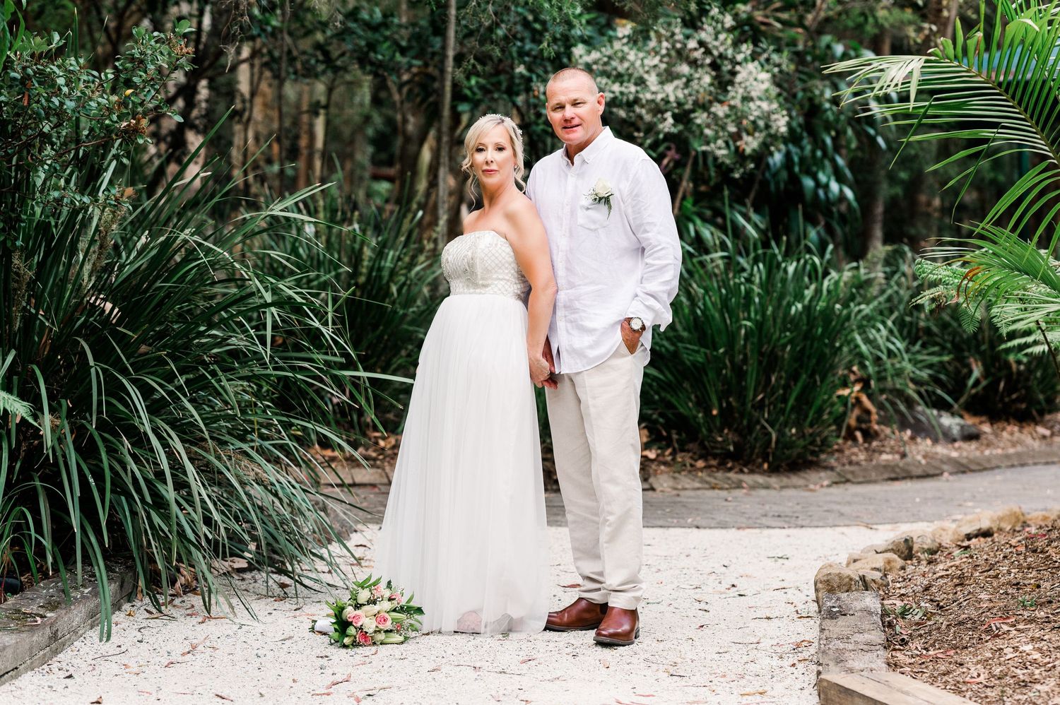 A wedding couple poses on a garden path surrounded by lush tropical plants and greenery.
