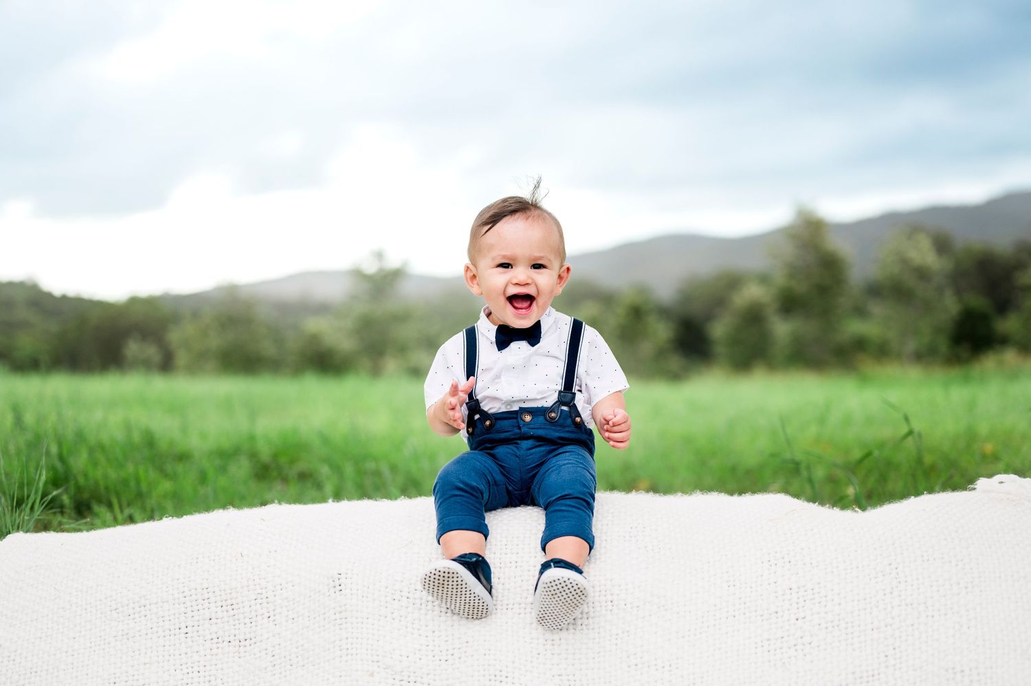 Happy baby in suspenders and bow tie sits on white blanket in grassy field with mountains behind.