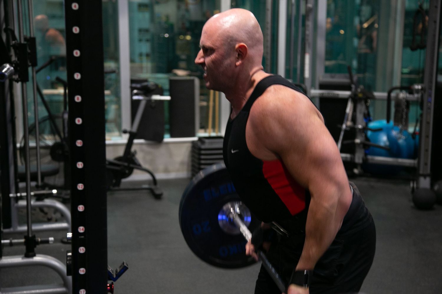 Bald muscular man in black tank top performing bent-over barbell rows in a dark gym.