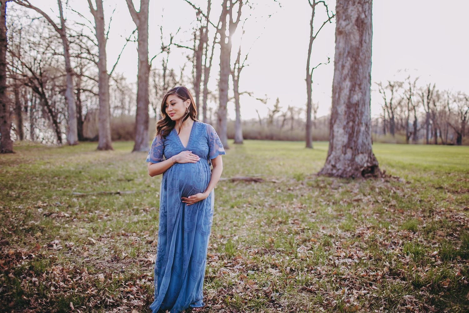 A pregnant woman in a flowing blue dress poses in a serene wooded setting.