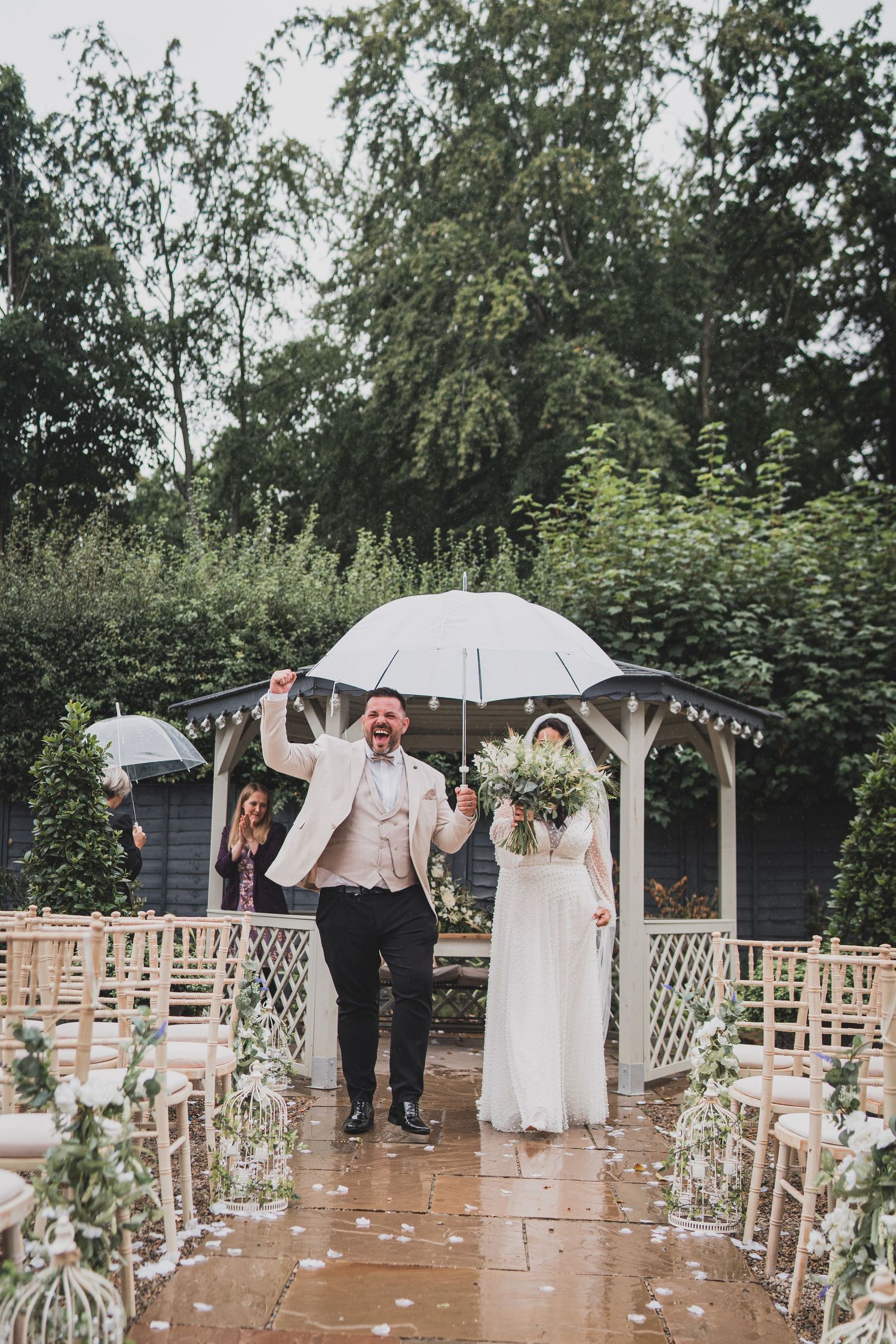 A joyful moment in the rain under a white umbrella on a garden wedding aisle decorated with flowers and lanterns.
