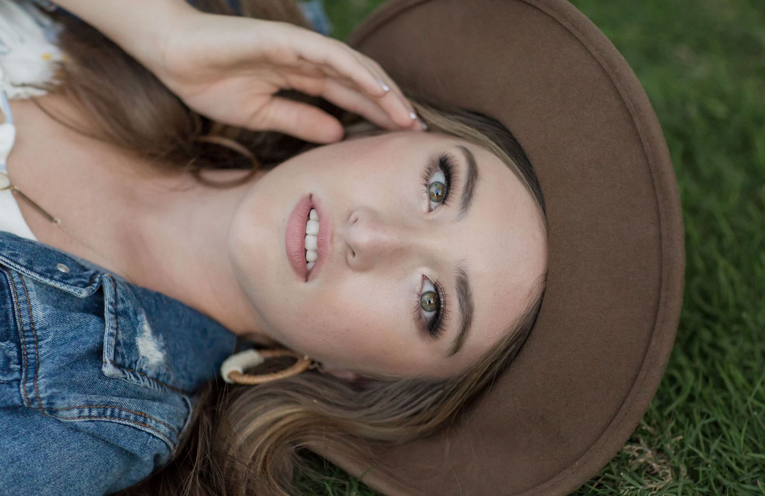 Close-up of young woman lying on grass wearing brown wide-brim hat, smiling at camera.