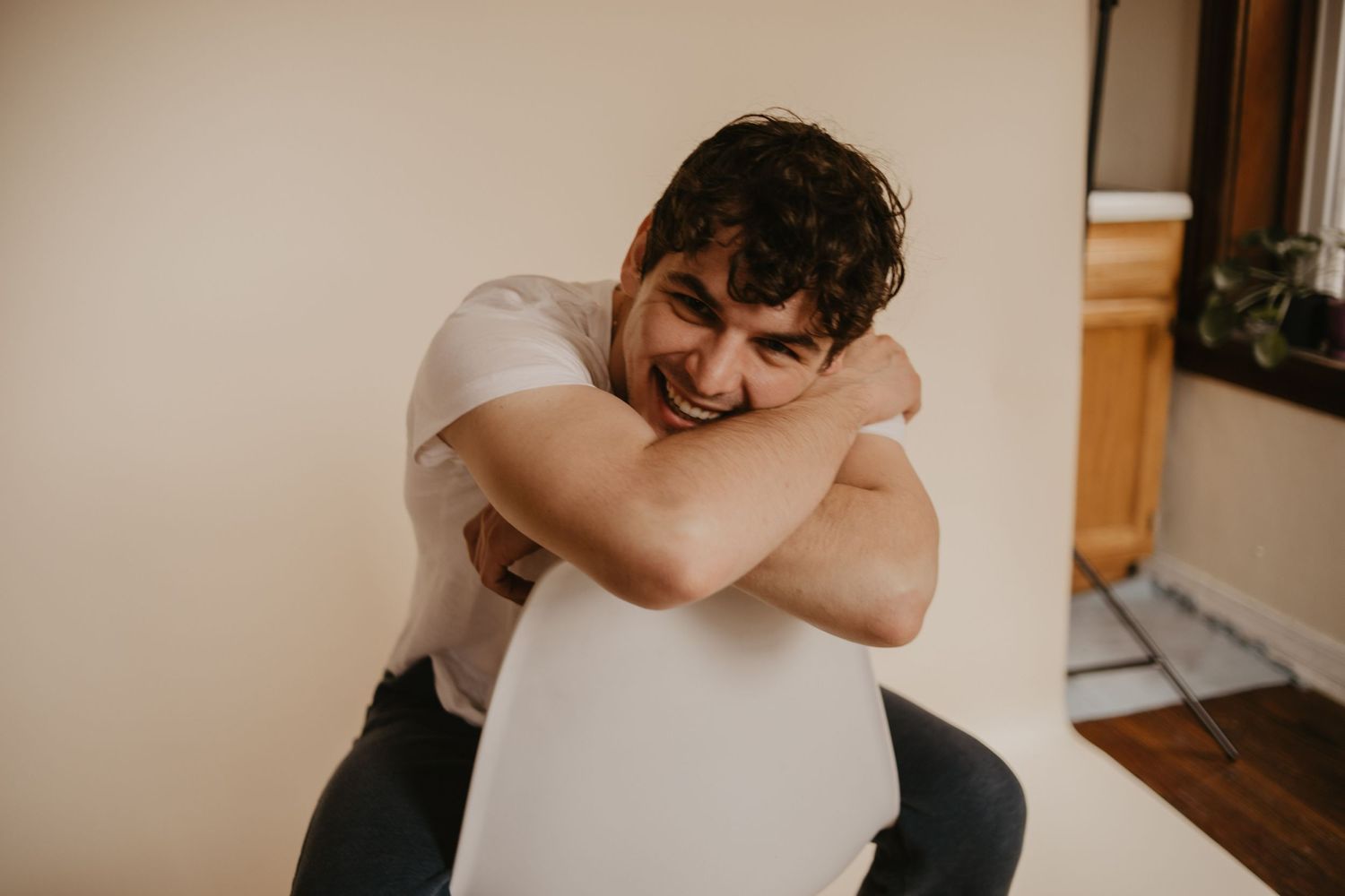 Person in white t-shirt hugging their knees while sitting against a beige wall.