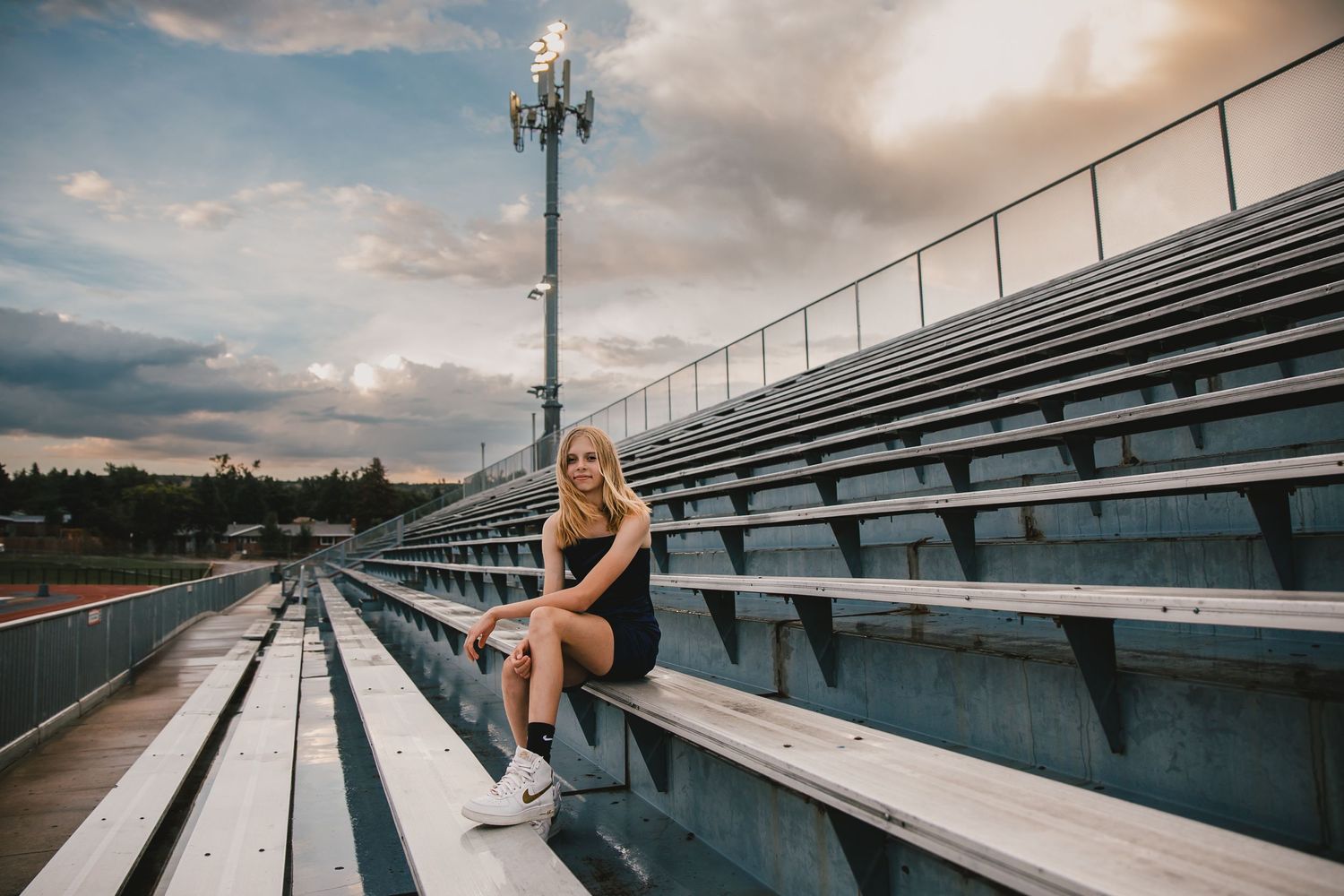 Athletic woman sits on stadium bleachers at sunset with floodlights and cloudy sky in background.