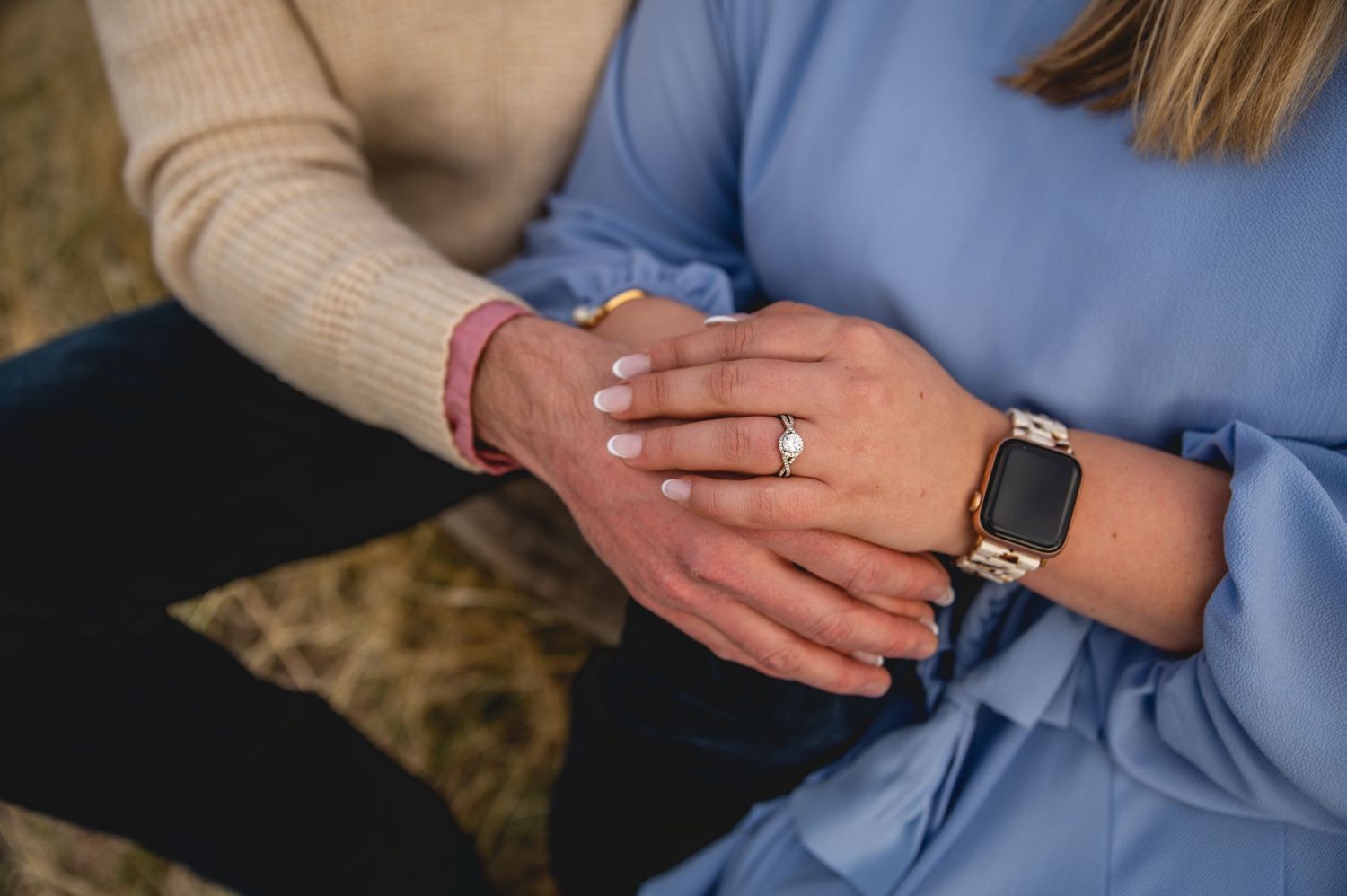 Close-up shot of hands wearing an engagement ring and Apple Watch against casual clothing.