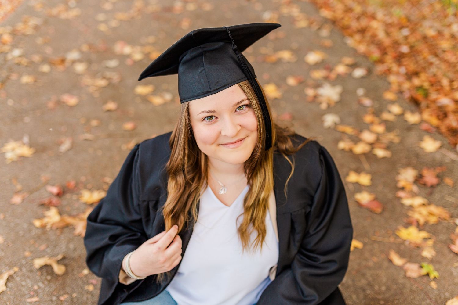 A graduate in academic regalia poses outdoors among fallen autumn leaves.
