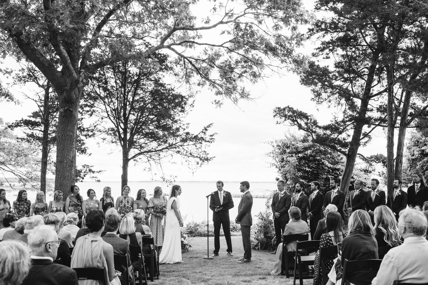 A panoramic view of wedding guests gathered outdoors under tall trees during a ceremony.