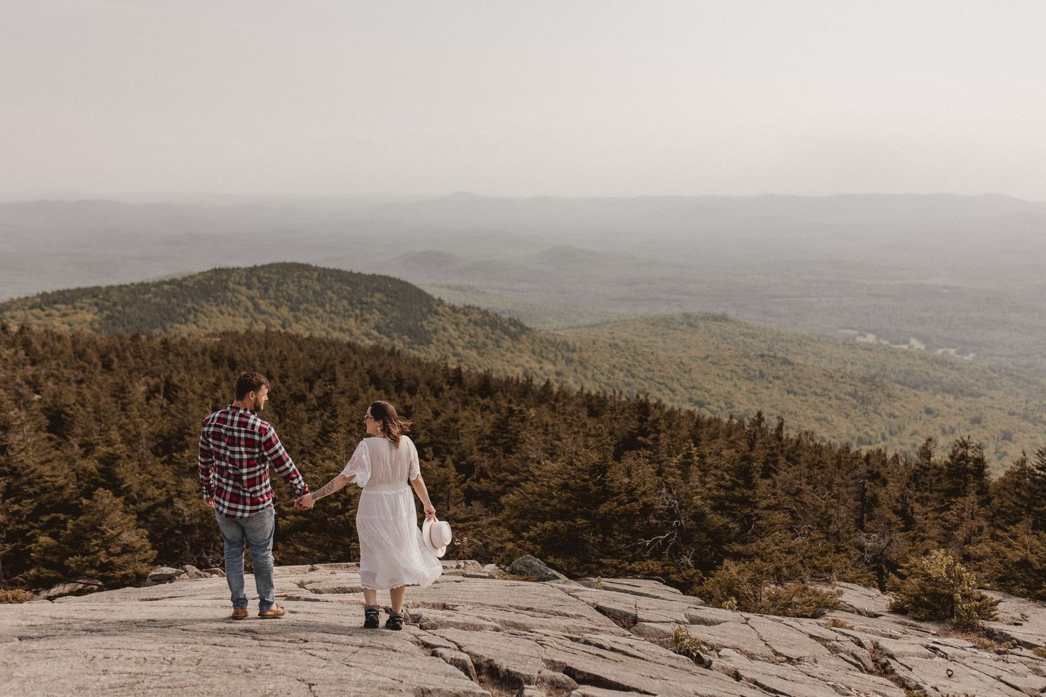 Couple holding hands while standing on rocky mountain summit overlooking vast landscape.