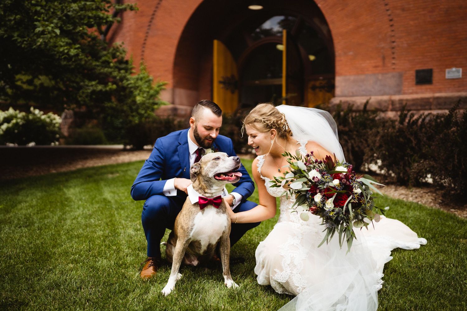 Couple shares moment with their dog on grassy lawn outside brick building.
