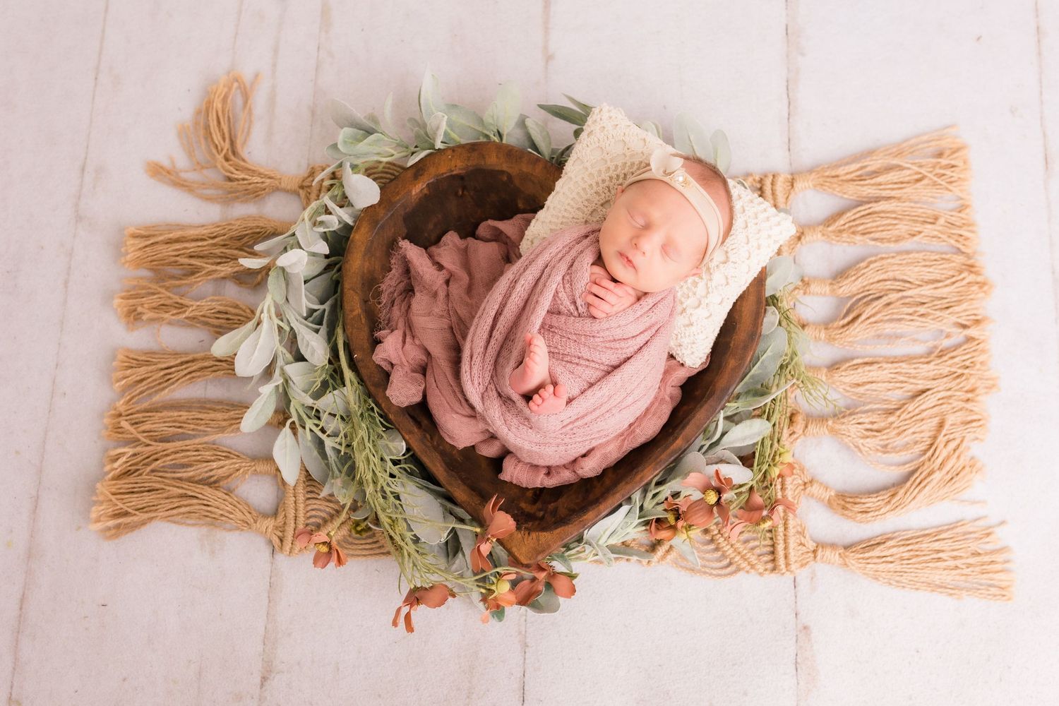 Sleeping newborn baby swaddled in pink wrap nestled in a wooden bowl on a boho fringe mat with greenery.