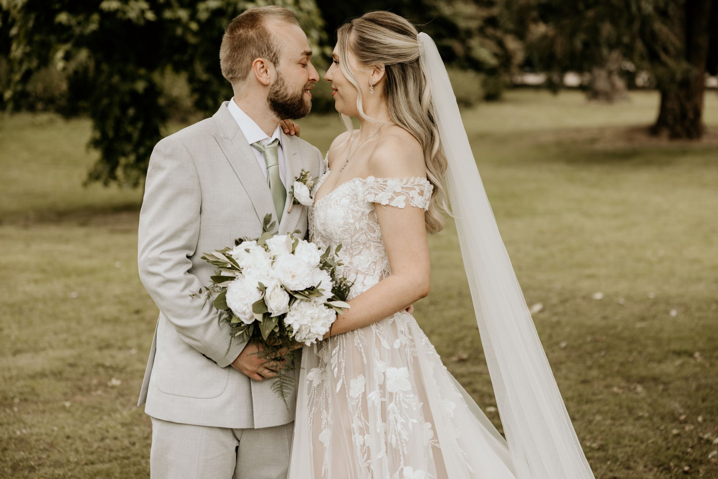 A romantic wedding photograph showcasing a couple kissing while holding a white floral bouquet in an outdoor garden setting.