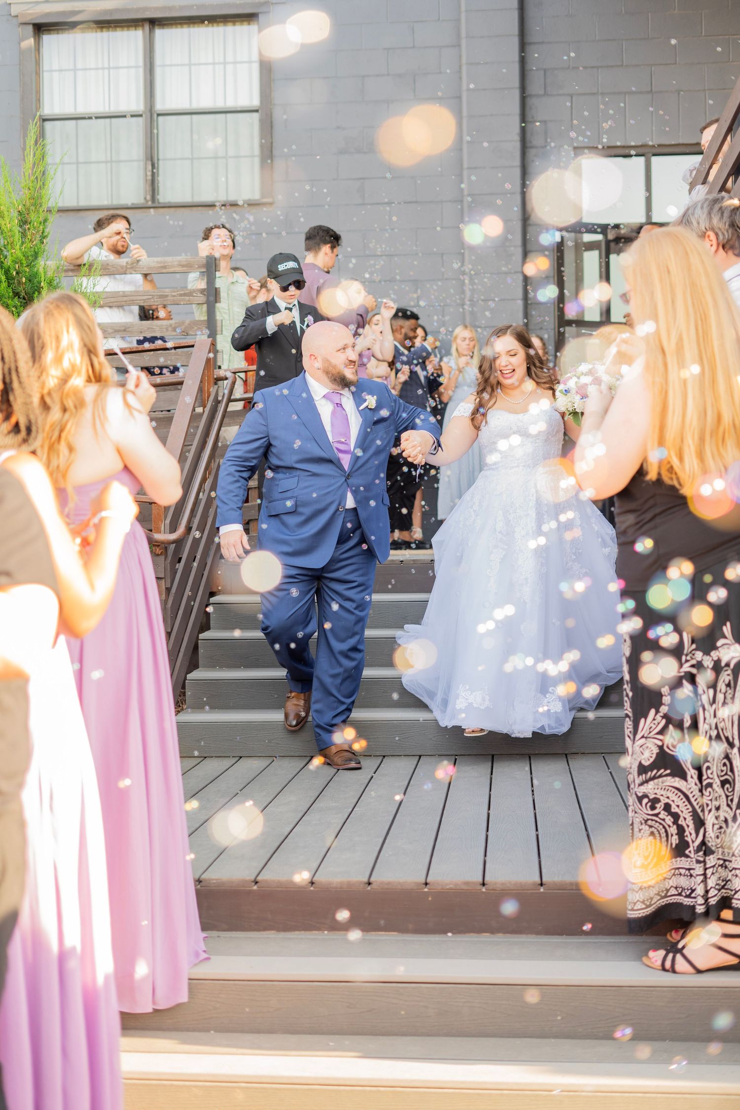Newlyweds exit through a joyful bubble sendoff, bride in blue gown and groom in navy suit, surrounded by cheering guests.