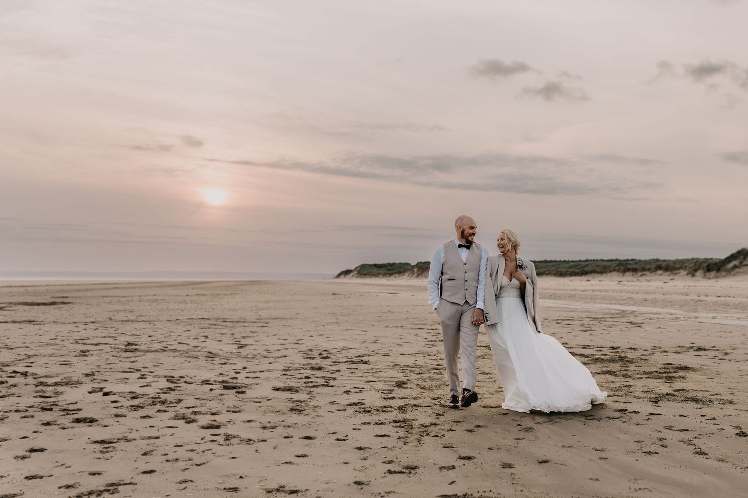 Newlyweds walking along a beach at sunset in formal wedding attire.