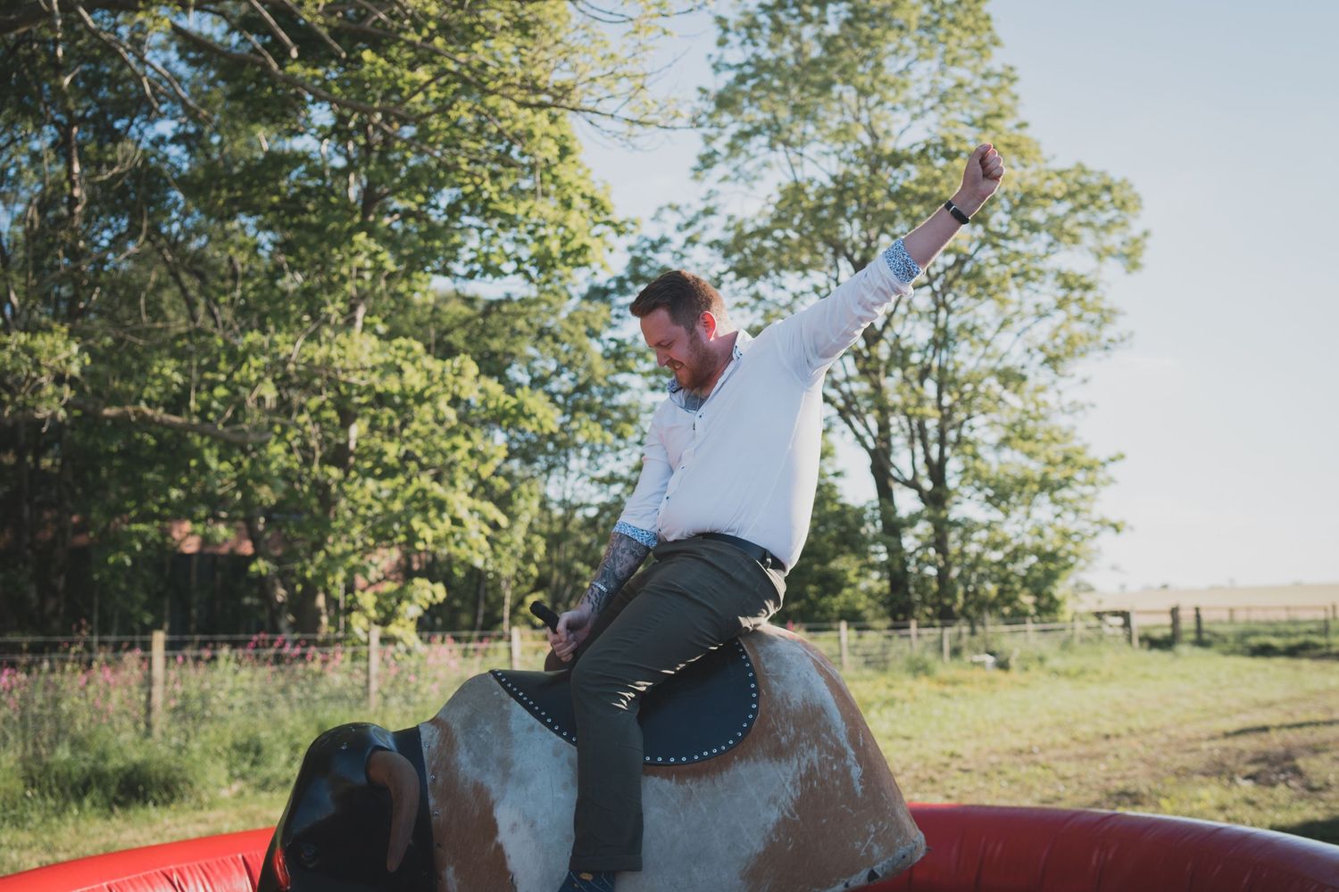 A person rides a mechanical bull outdoors with arms raised in excitement.