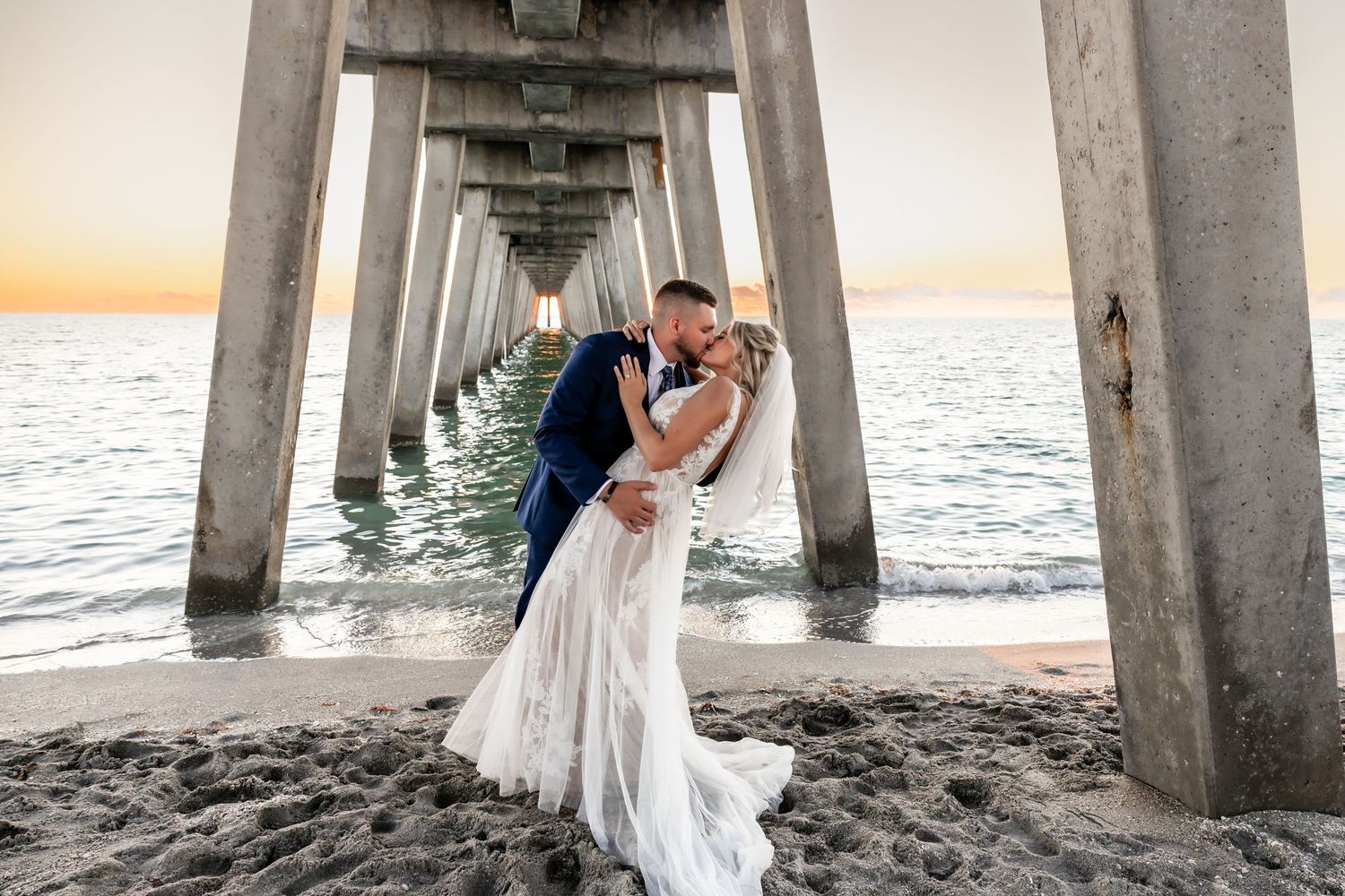 Bride and groom kissing under a pier at sunset on a sandy beach.