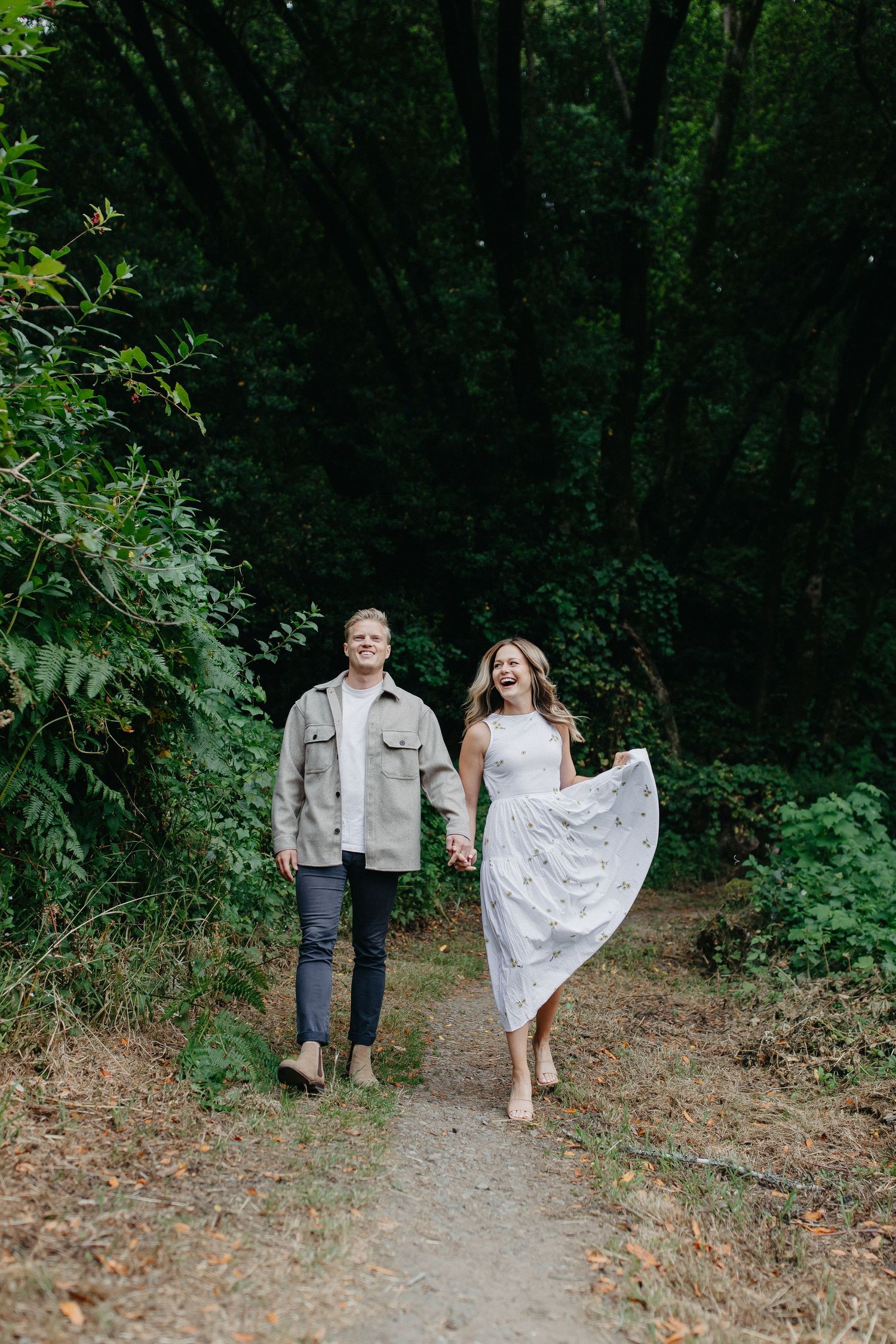 A couple walking together on a forest path as she playfully lifts her white dress.
