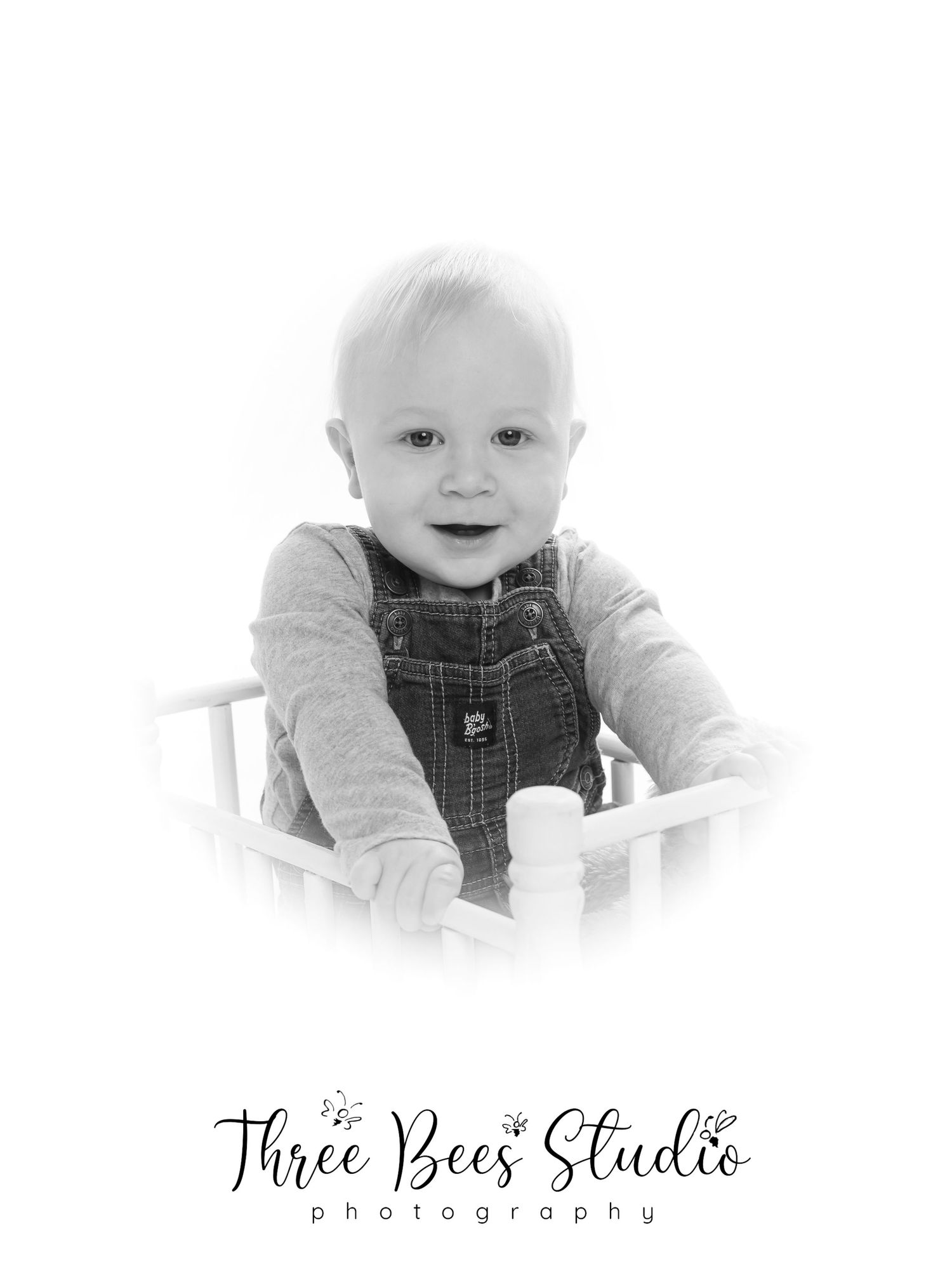 Professional black and white studio portrait of a smiling baby in overalls sitting in a white chair.