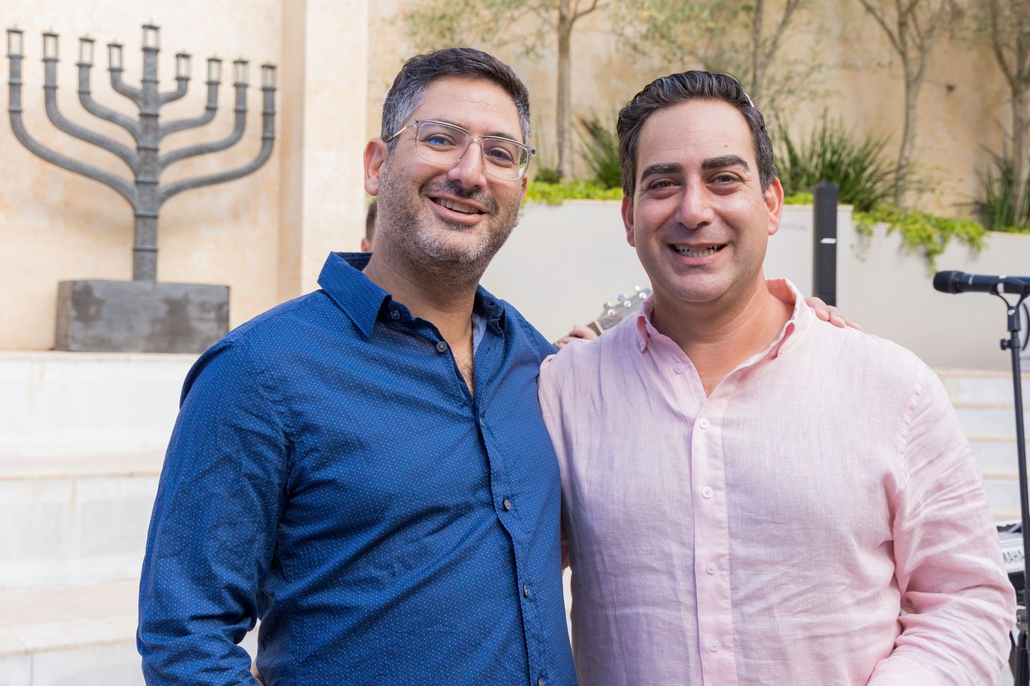 Two men smiling together in front of a large menorah decoration.