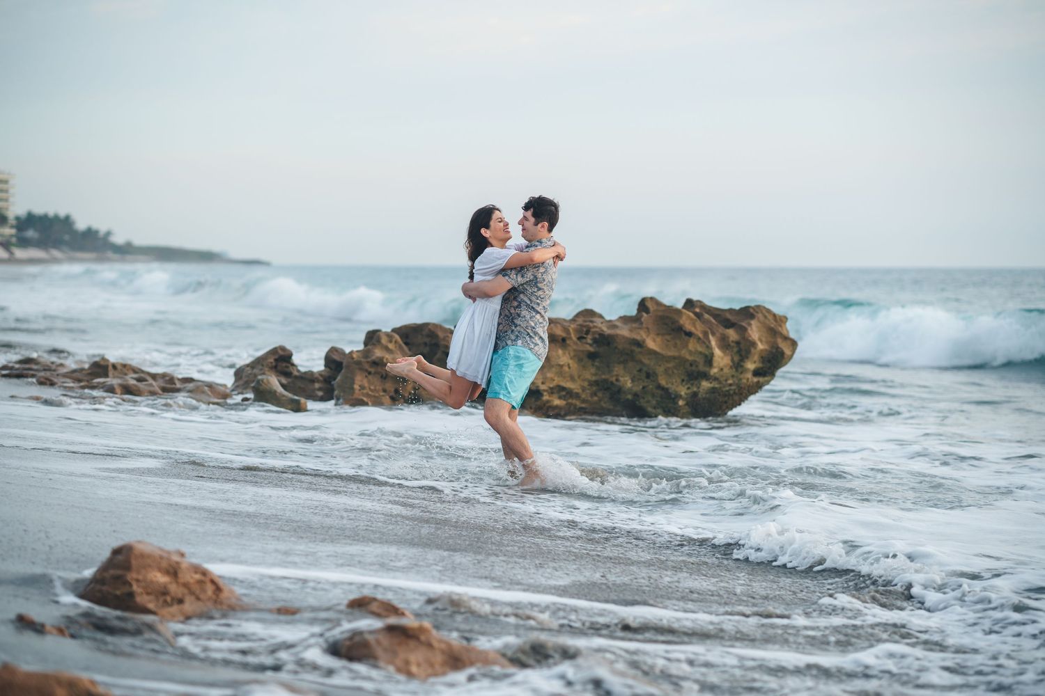 Romantic couple embracing on rocky beach with ocean waves in background.