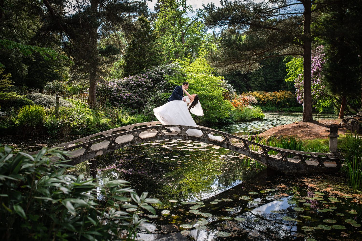 Romantic scene of a couple in white crossing an arched bridge over a pond in a lush garden setting.