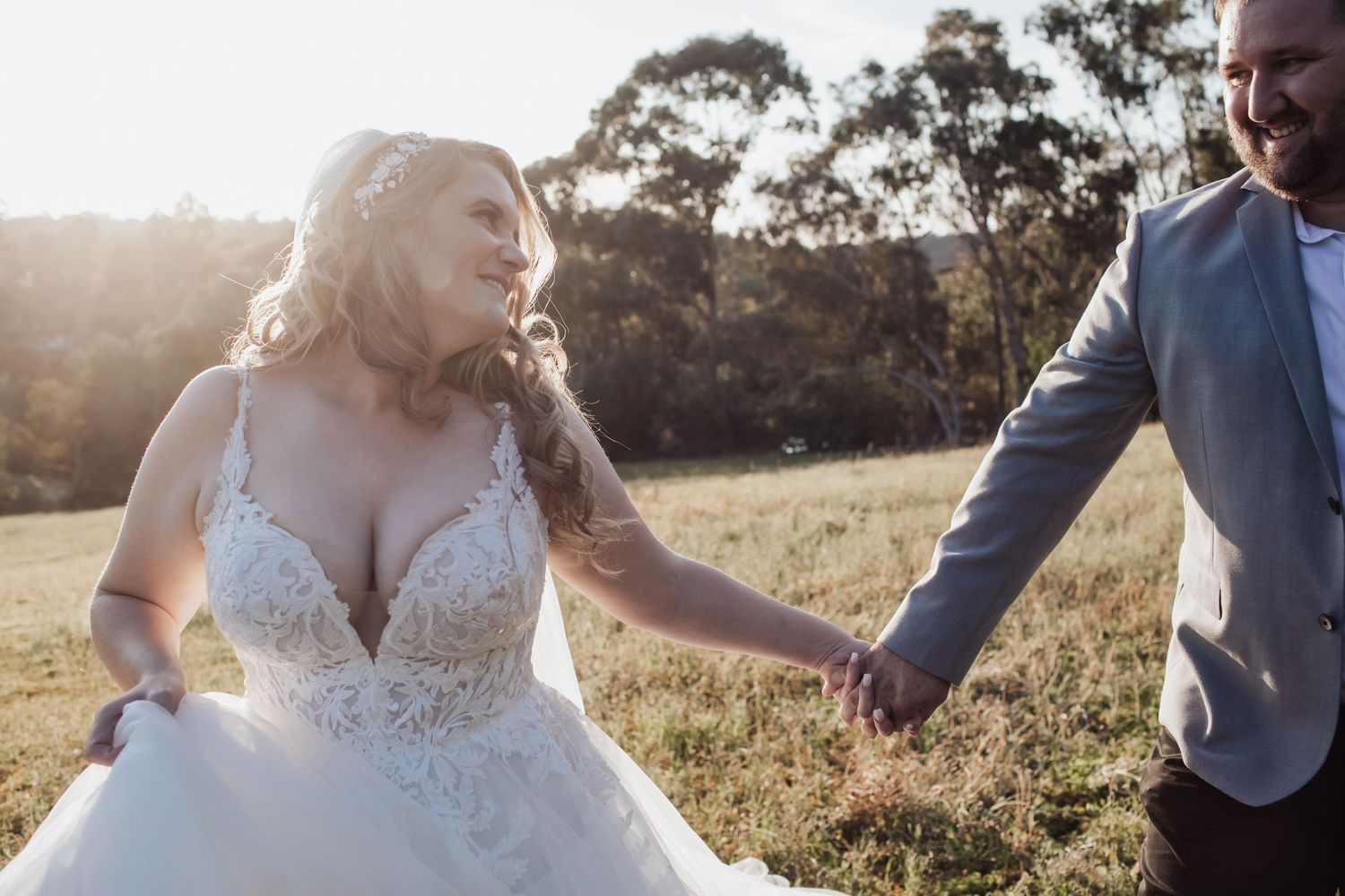 Couple walking through a sunlit field in wedding attire during golden hour.