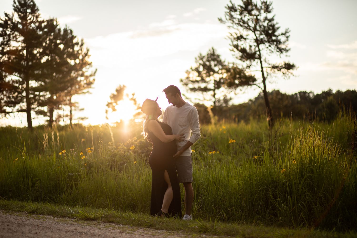 Couple embracing on path at sunset with tall pines and grass in background.