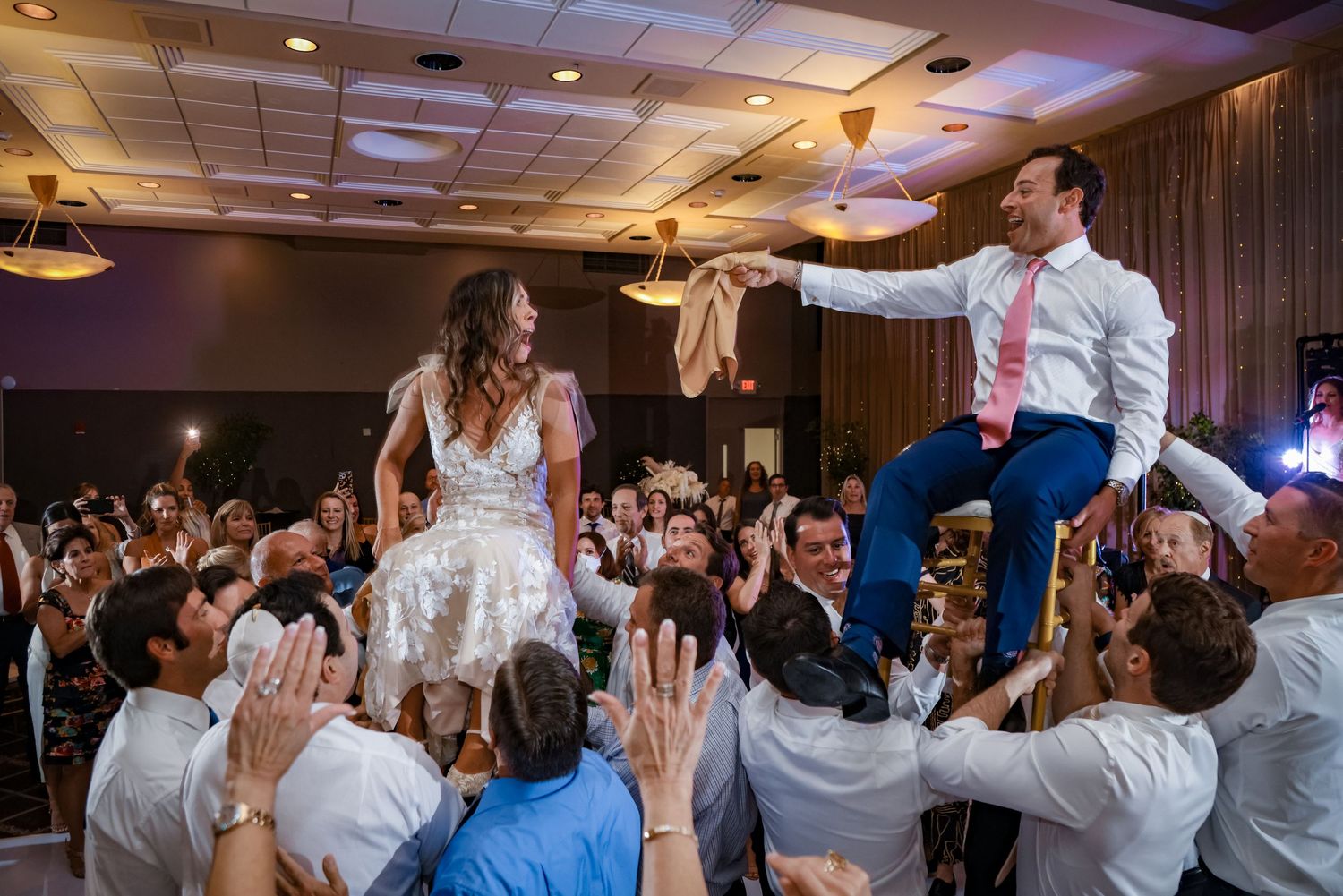 Wedding celebration with guests lifting couple during traditional Jewish hora dance.