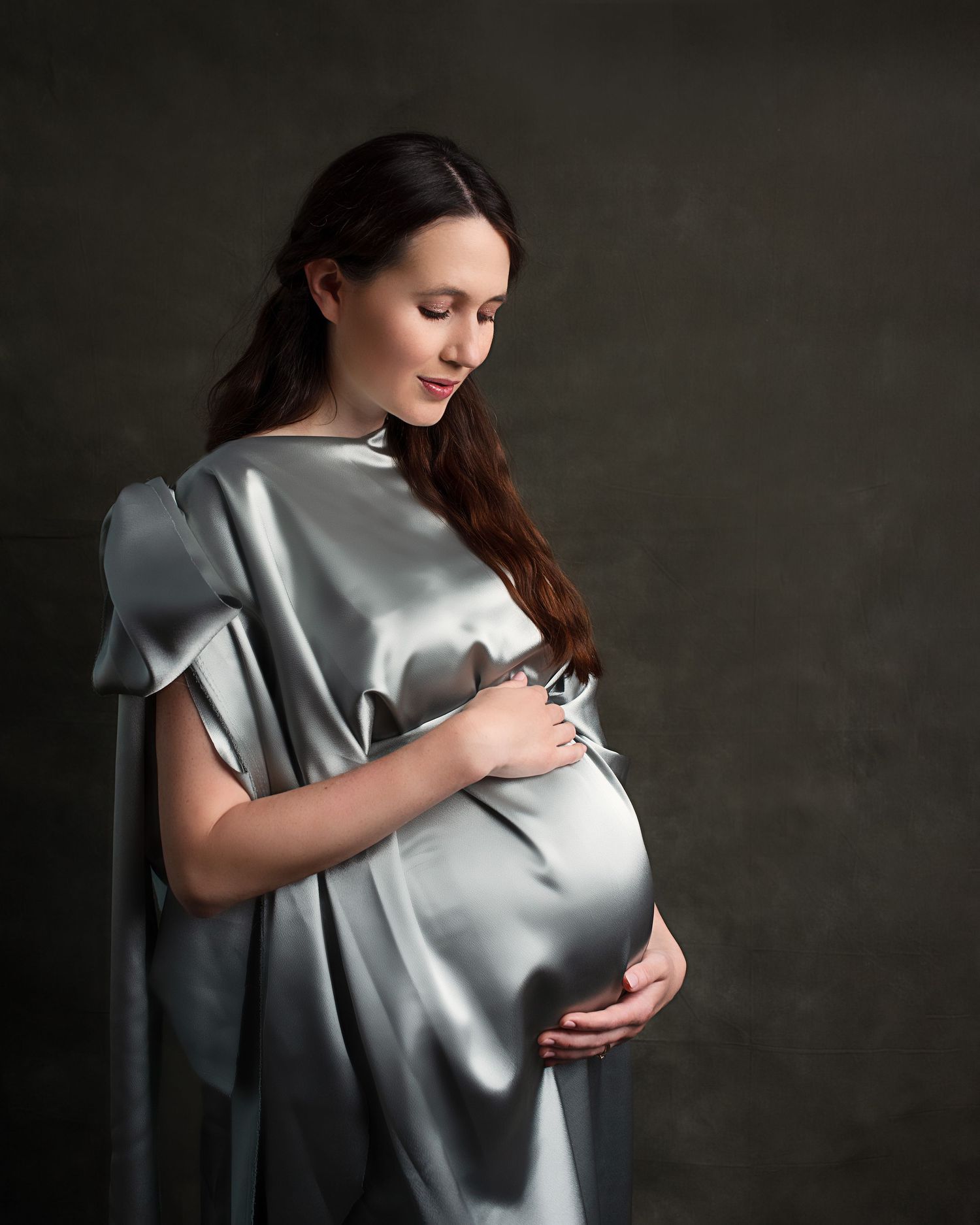 Pregnant woman in silver satin gown cradling baby bump against dark studio background.