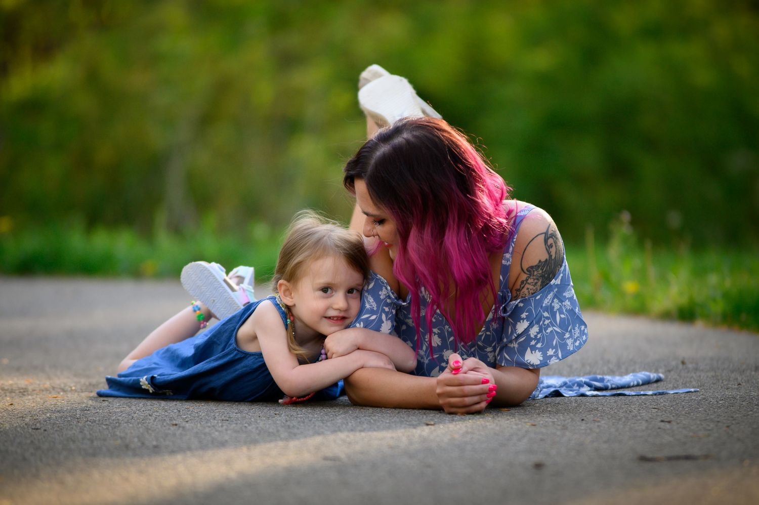 Mother with pink hair and tattoos lies on road smiling tenderly at young blonde daughter outdoors.