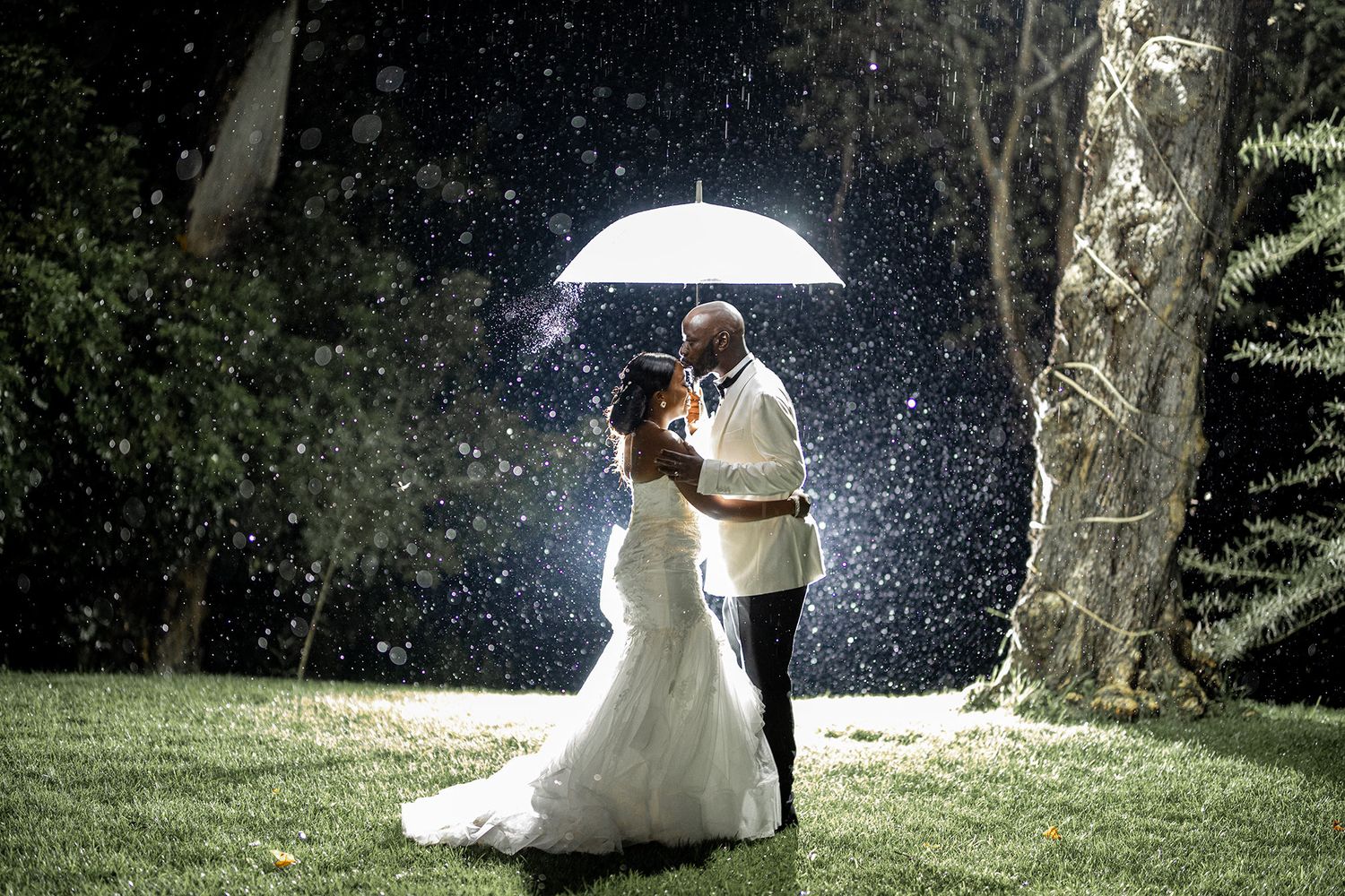 Couple shares romantic moment under white umbrella in the rain during wedding photoshoot.