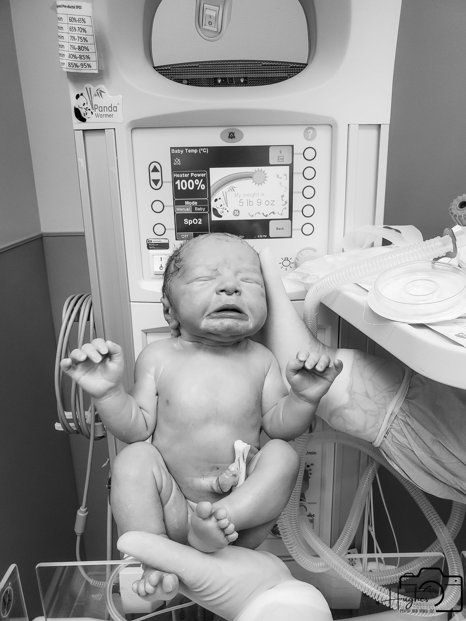 A newborn baby lies on a hospital bed with medical equipment in the background.