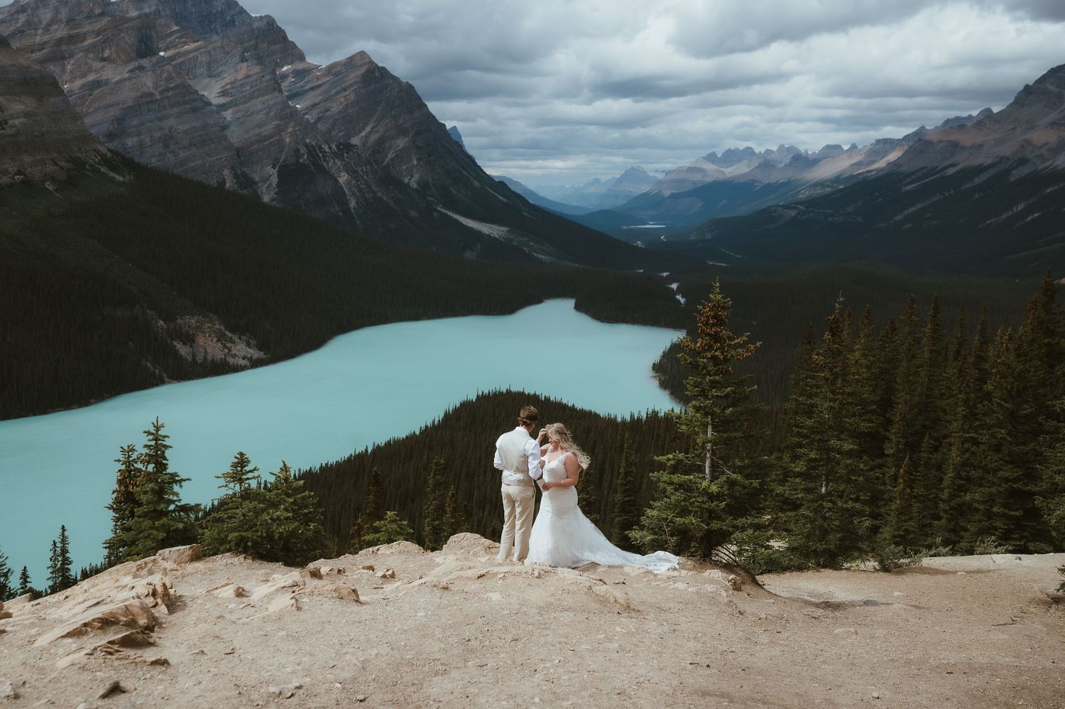 Bride and groom standing on mountain overlook with turquoise lake and evergreen forest below.