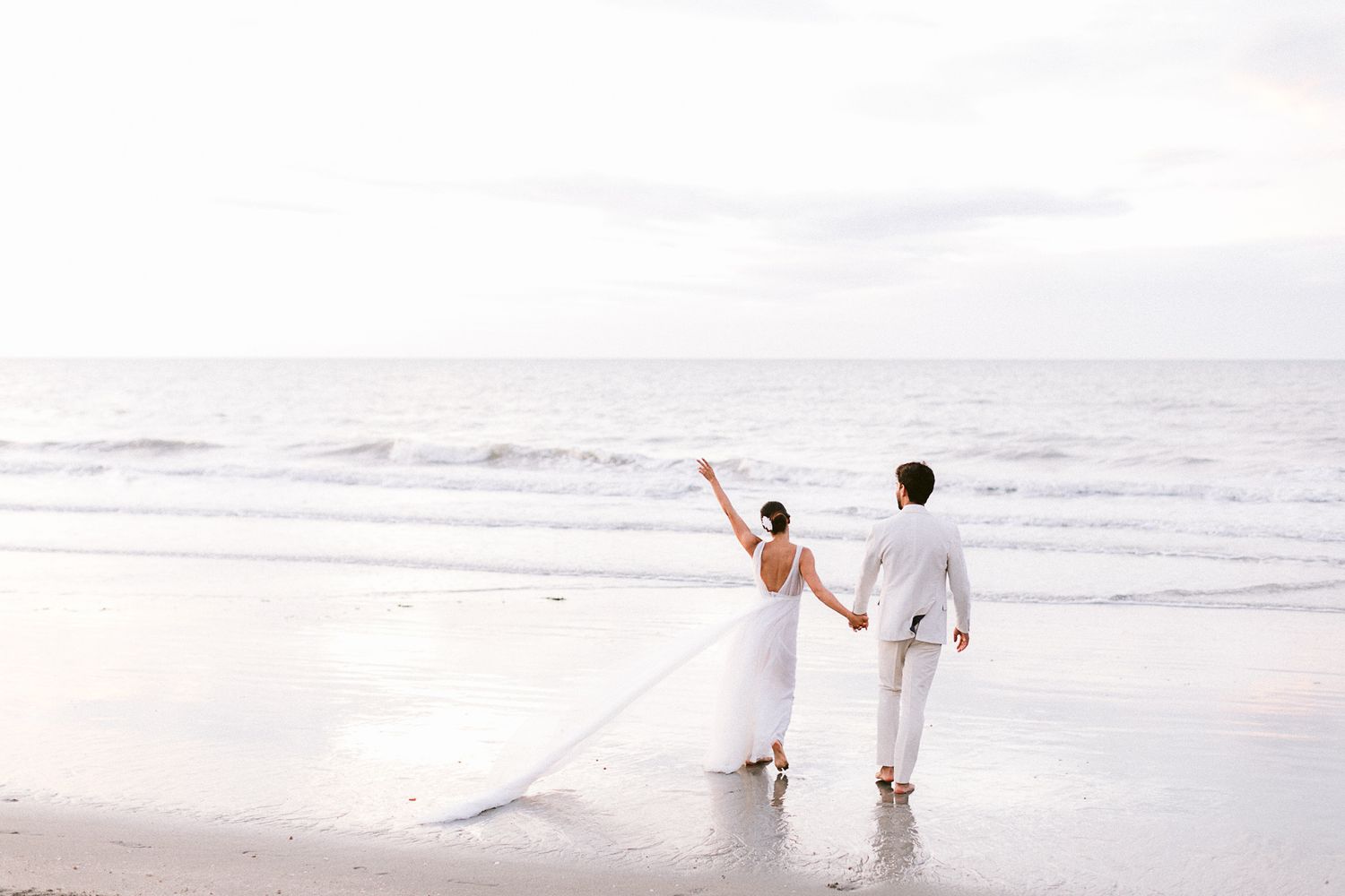 A couple in white attire walks along a misty beach shore at sunset.