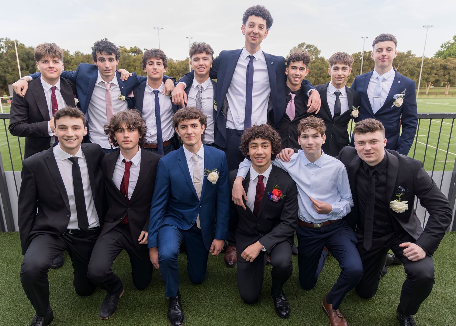 Group photo of young men in formal attire posing together outdoors on a sports field.