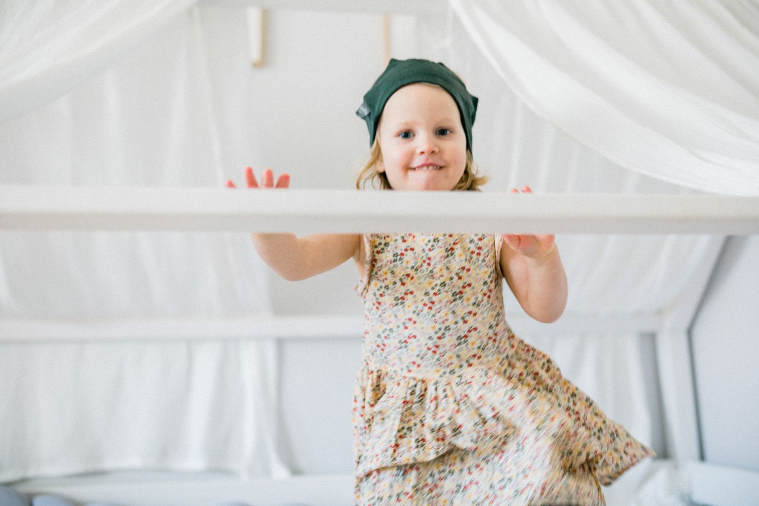 Young child in floral dress and turquoise hat playing beneath white drapes.