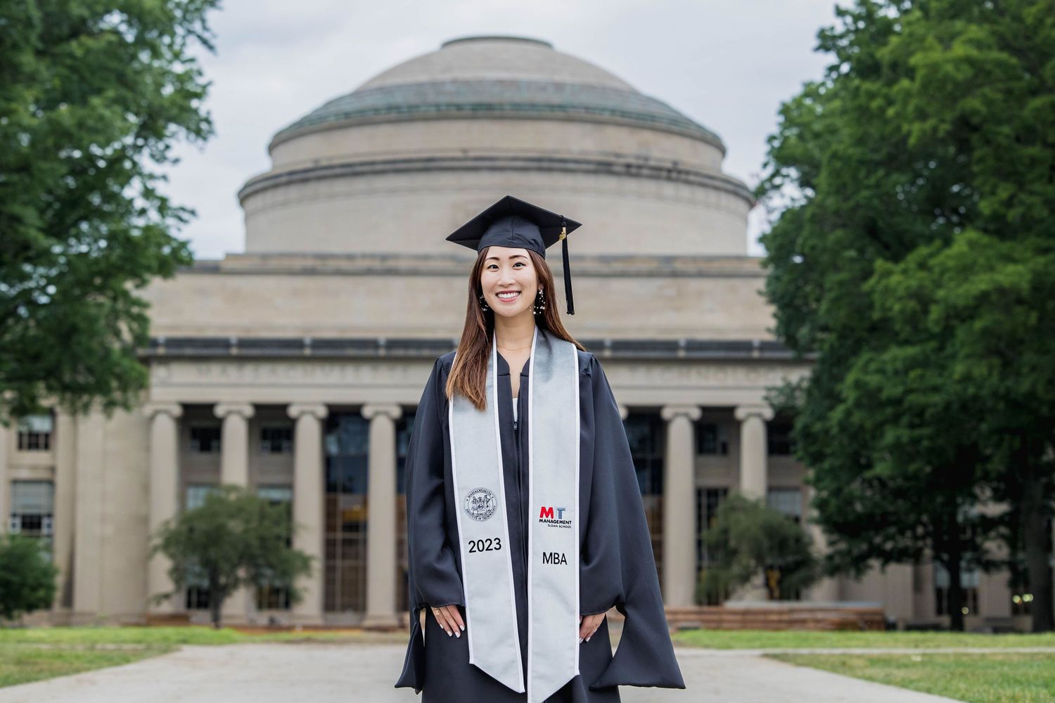 Graduate in cap and gown standing in front of MIT's iconic dome building.