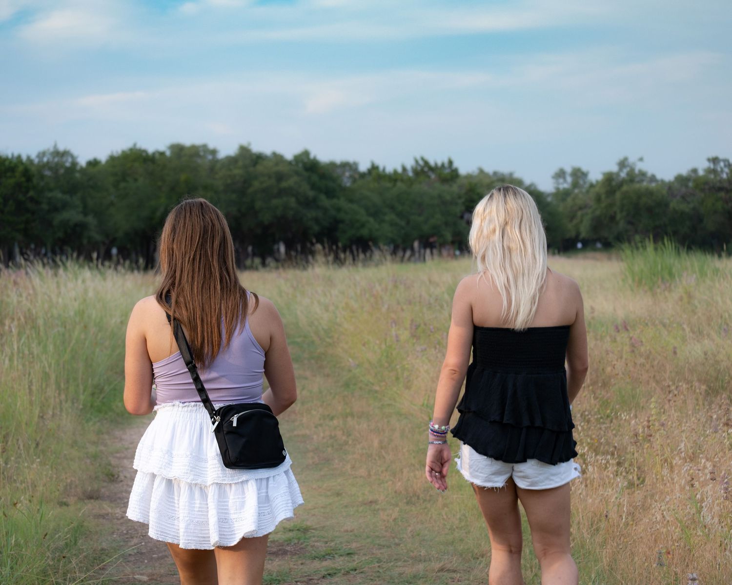 Two young women walking down a dirt path through green fields, viewed from behind.