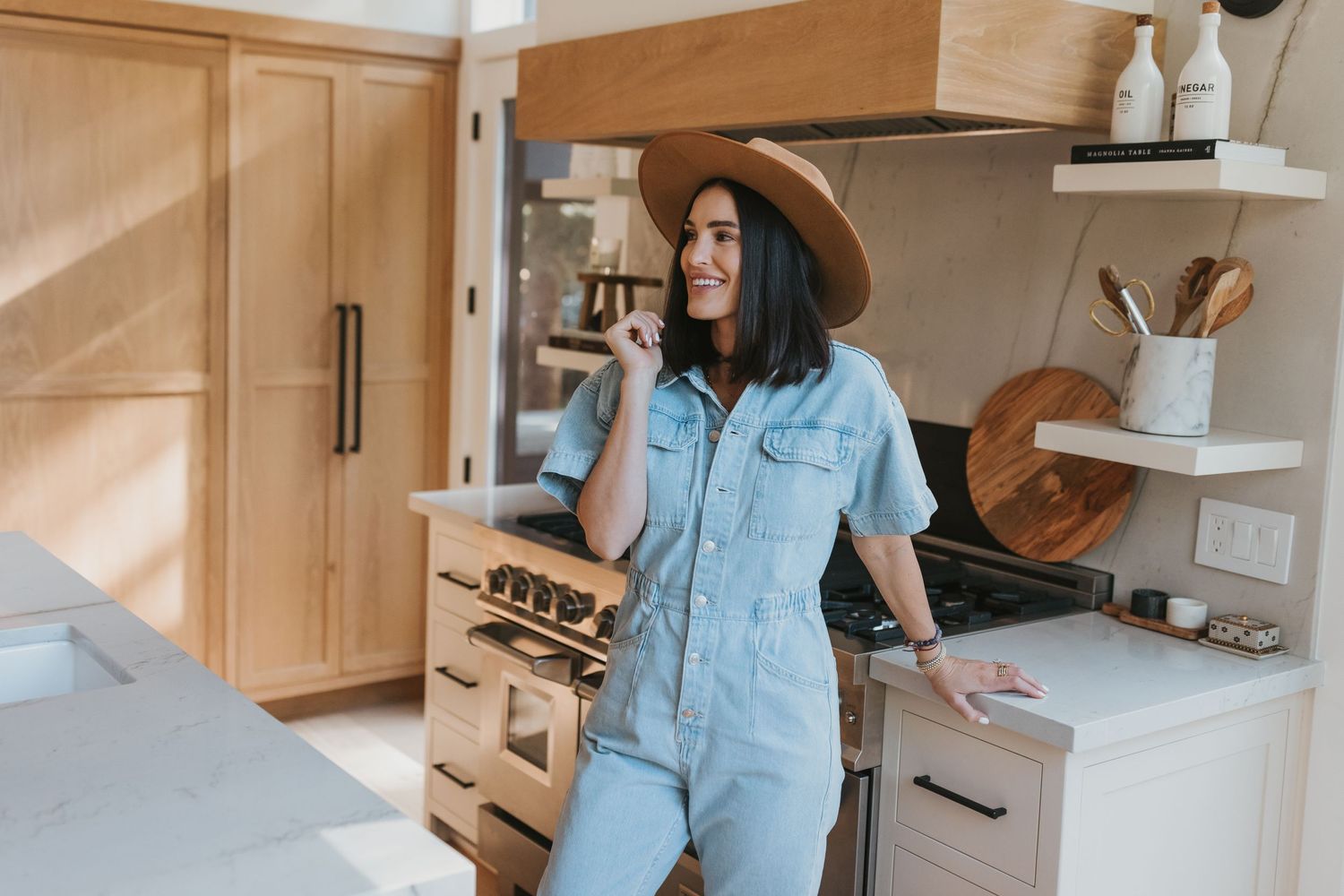 Casual scene of denim jumpsuit and wide brim hat in modern farmhouse kitchen with light wood cabinets.