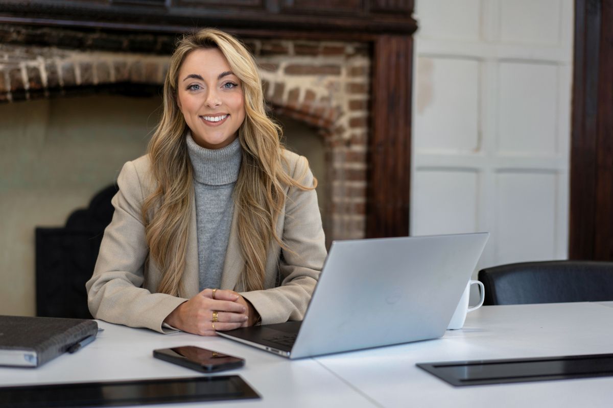 Woman in grey turtleneck and beige blazer smiles while working on laptop at desk.